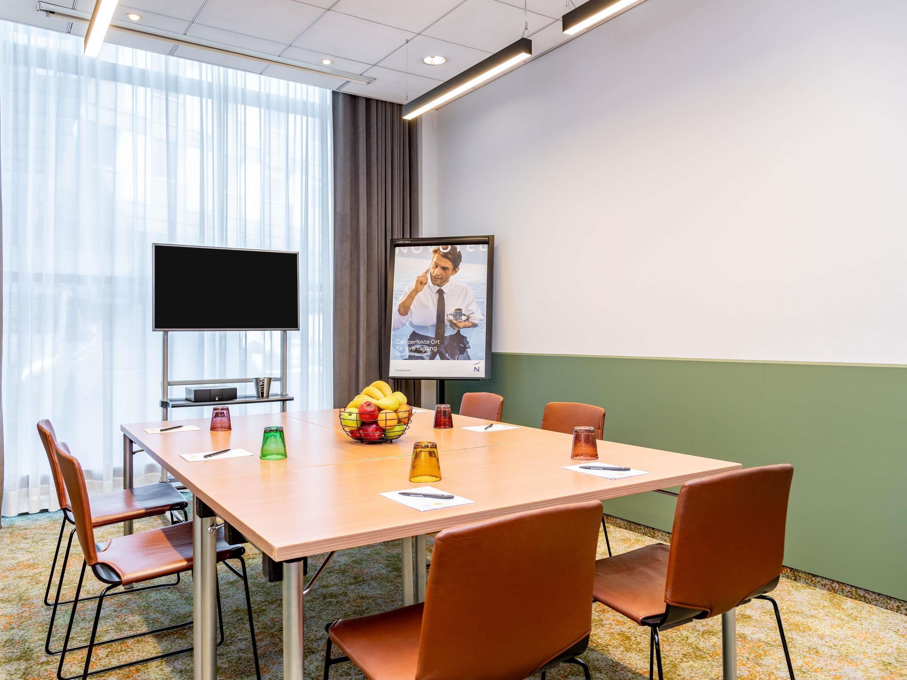 A conference room with a long table, six chairs, a TV screen, a poster, and a fruit bowl.
