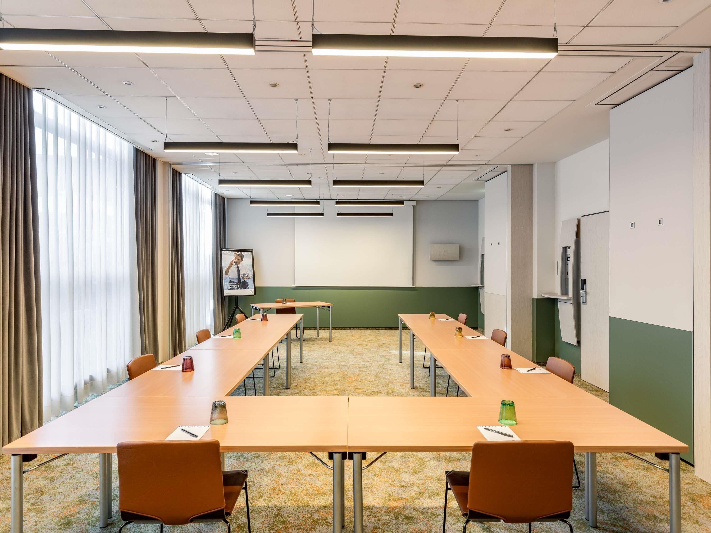 A conference room with long tables, chairs, a projector screen, and a large window with curtains.