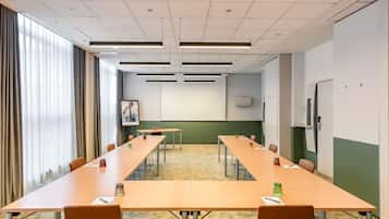 Un salón de conferencias con mesas largas, sillas, una pantalla de proyector y un gran ventanal con cortinas.