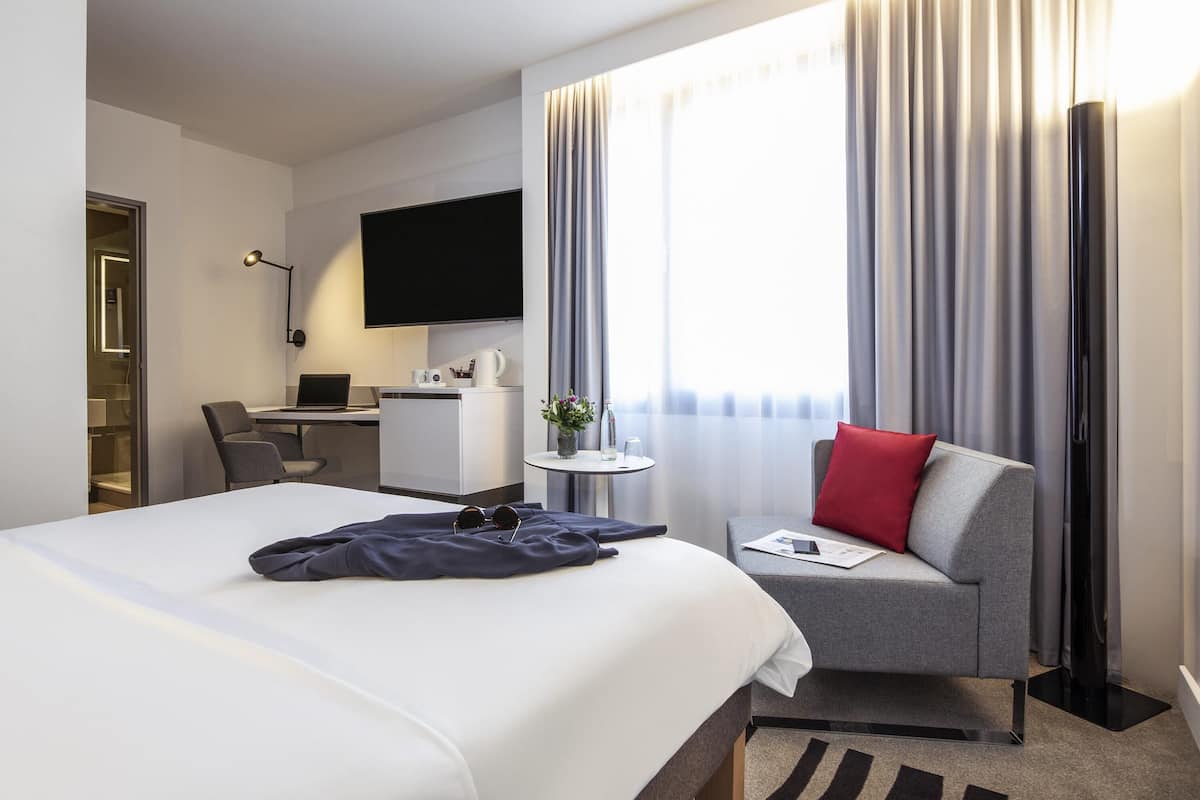 Une chambre d’hôtel avec un lit, un bureau avec un ordinateur, une chaise, une petite table avec une plante et un téléviseur fixé au mur.