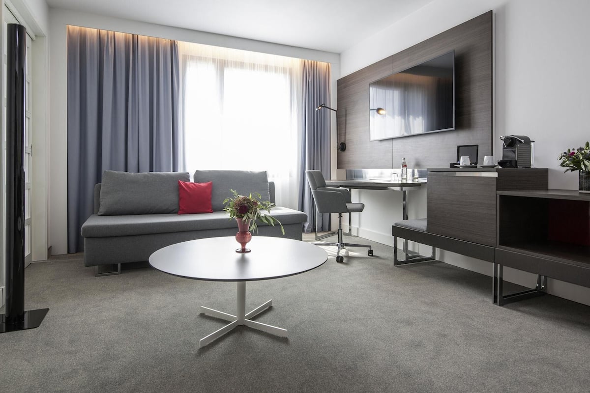 A modern living room with a sofa, a round table, a desk, and a flat-screen TV mounted on the wall.
