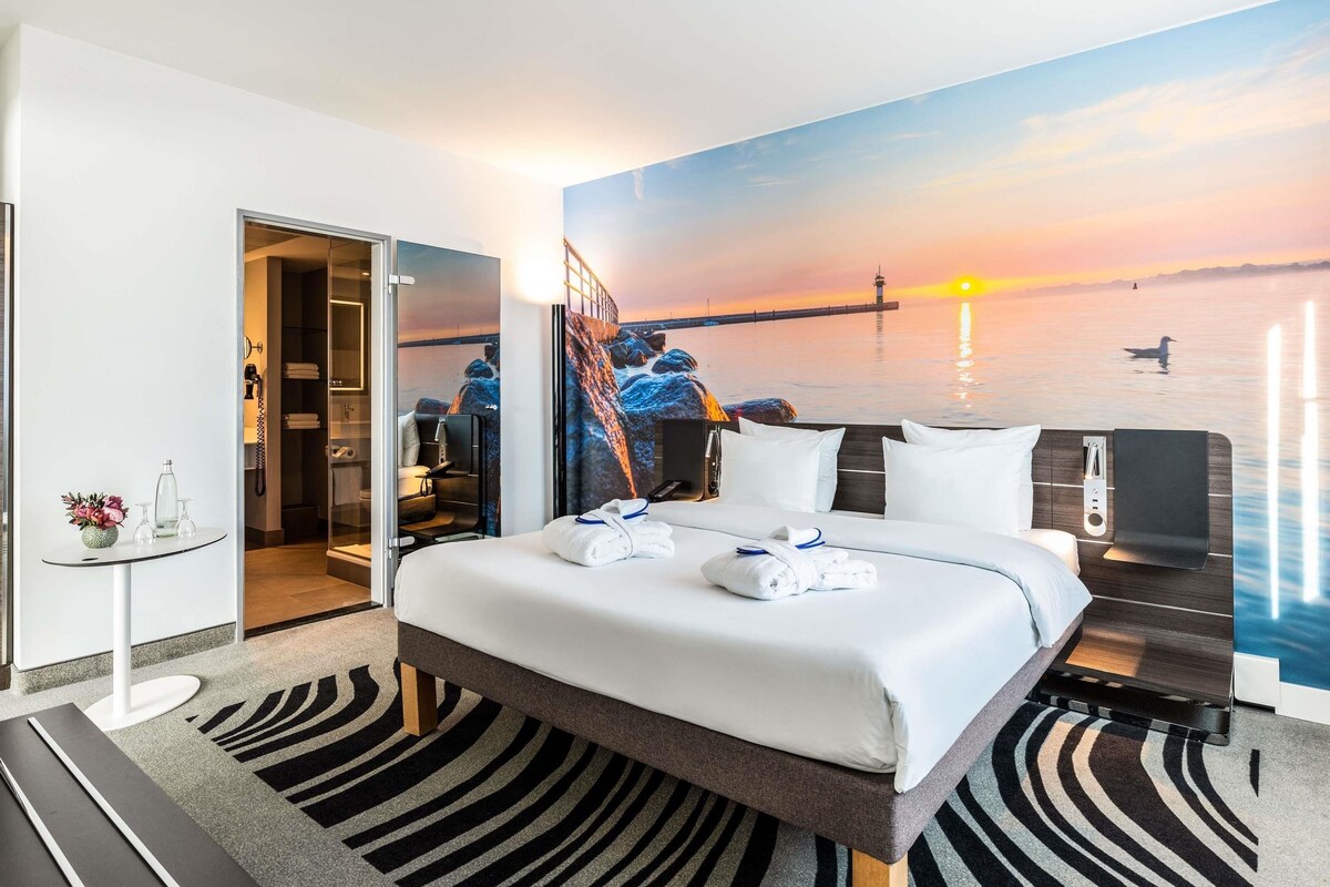 Een hotelkamer met een bed, een bureau, een stoel, een bijzettafeltje met een vaas en een schilderij van een zonsondergang boven het water.