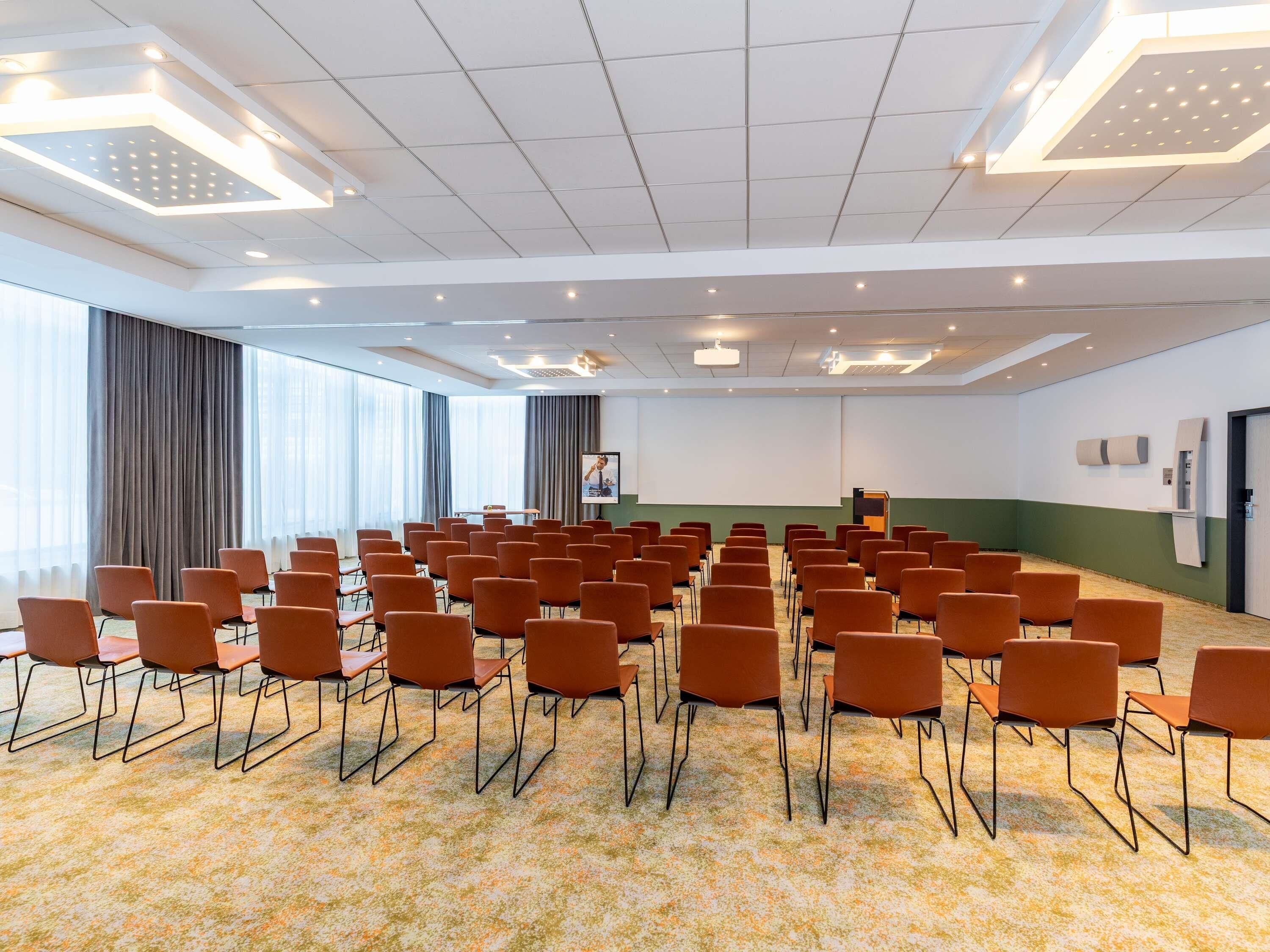 Ein Konferenzraum mit Reihen orangener Stühle, einem Rednerpult und einer großen Leinwand.