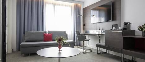 Un salon moderne avec un canapé gris, une table basse ronde, un téléviseur fixé au mur et un bureau avec une chaise.