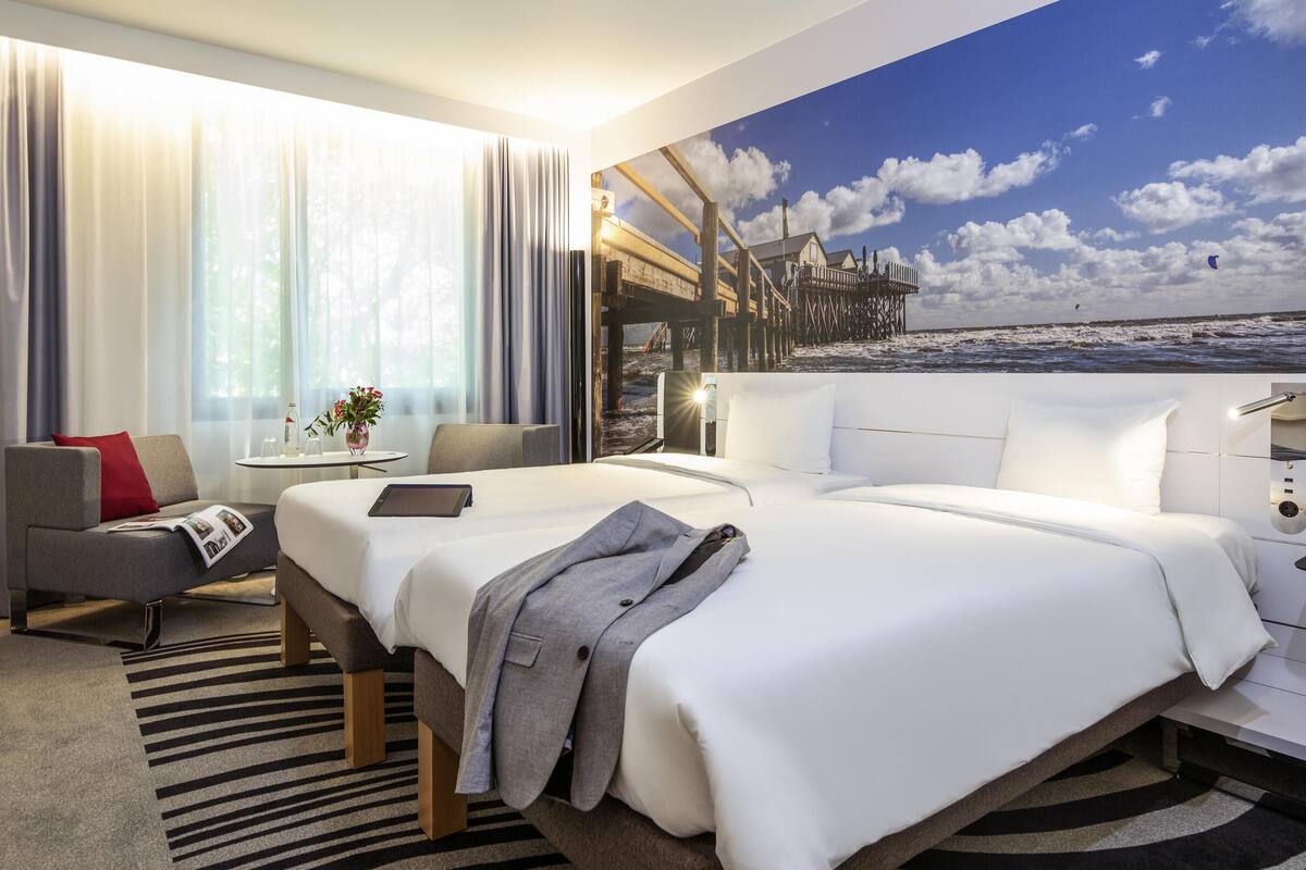 Ein Hotelzimmer mit einem großen Bett, einer Couch, einem kleinen Tisch mit einer Vase und einem Wandgemälde mit einer Strandansicht.