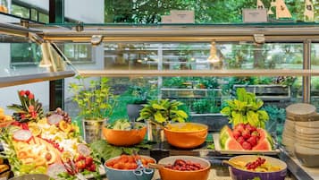 Ein Buffet mit verschiedenen Früchten, Gemüsesorten und Käse, mit Blick auf grüne Vegetation draußen.
