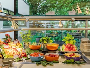 A buffet with various fruits, vegetables, and cheeses, with a view of greenery outside.