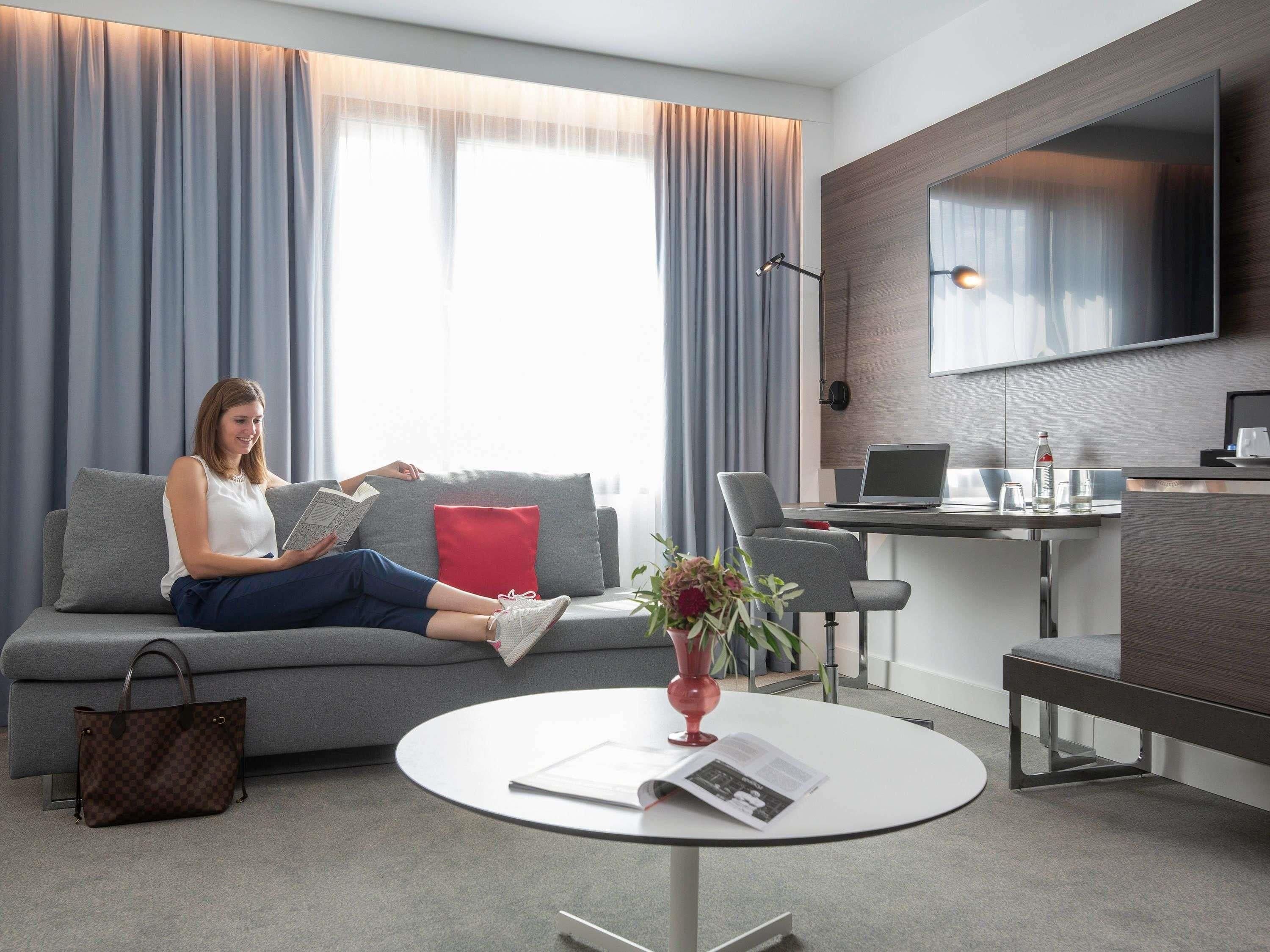 Quarto de hotel moderno com sofá cinza, mesa de centro redonda com uma revista, escrivaninha com laptop e uma TV grande fixada na parede.