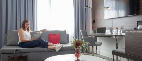 Quarto de hotel moderno com sofá cinza, mesa de centro redonda com uma revista, escrivaninha com laptop e uma TV grande fixada na parede.