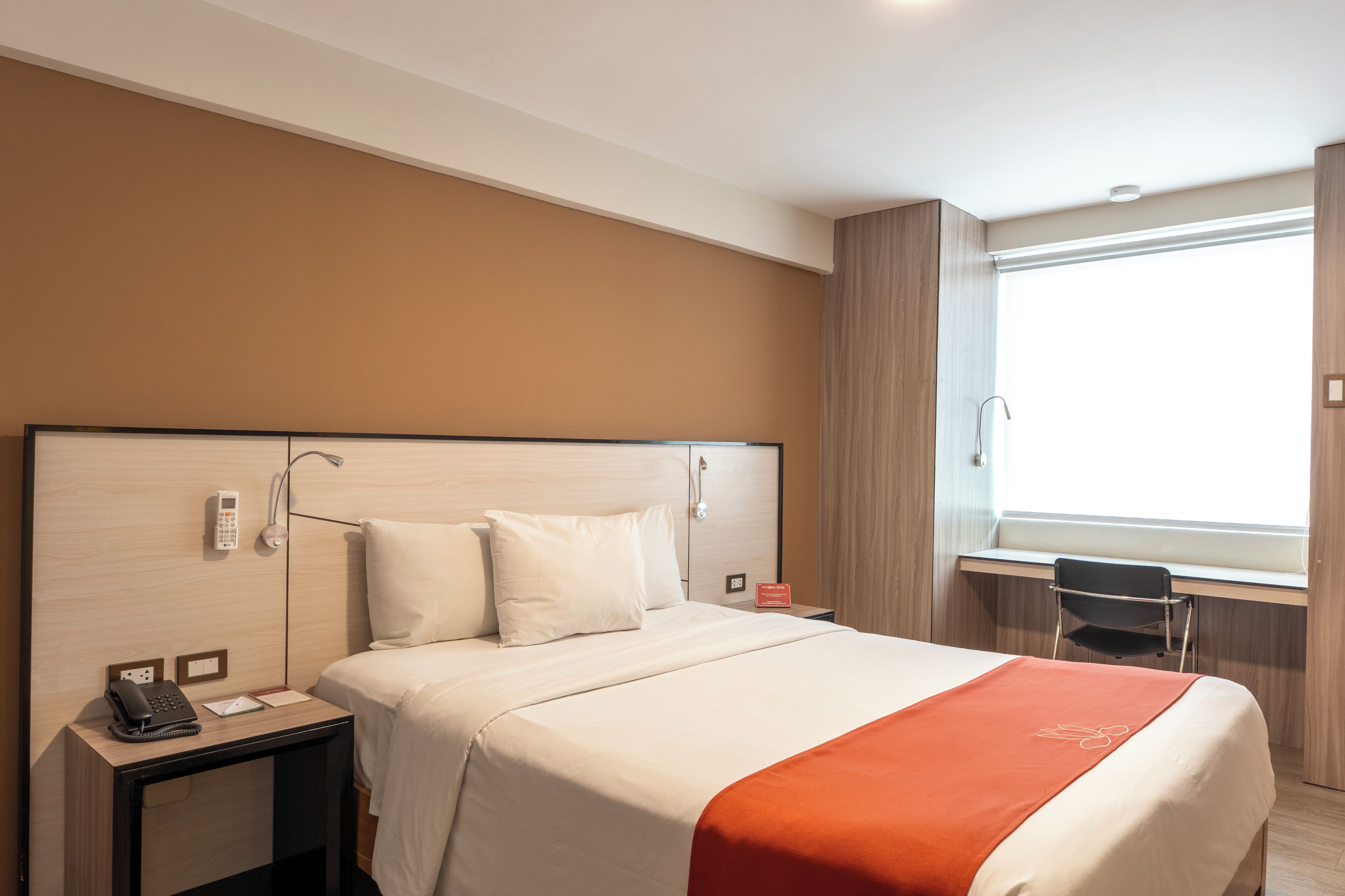 A hotel room with a large bed, a bedside table with a phone, a desk with a chair, and a window with curtains.