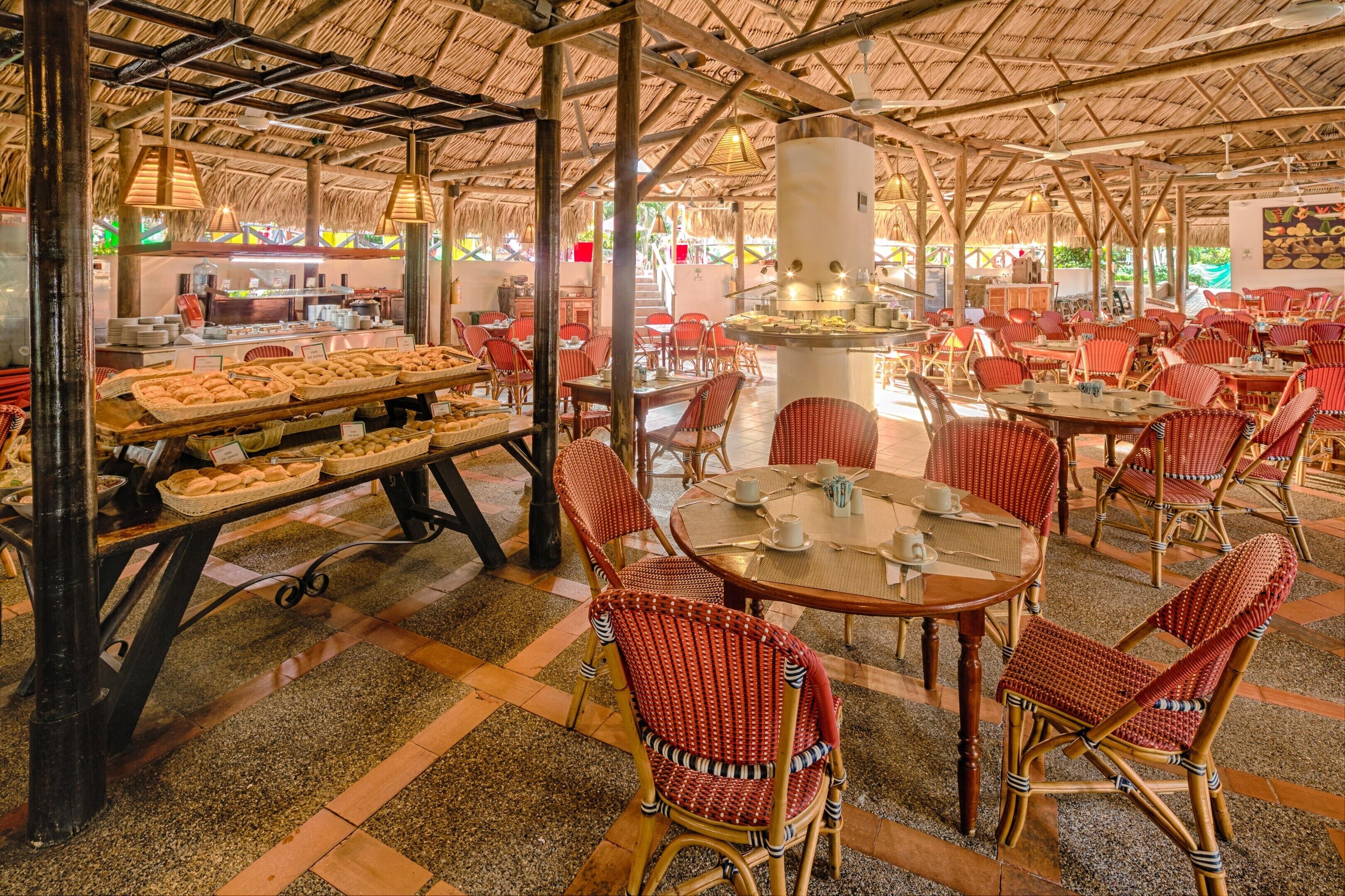 A restaurant with a thatched roof, wicker chairs, and a buffet area with bread and pastries.