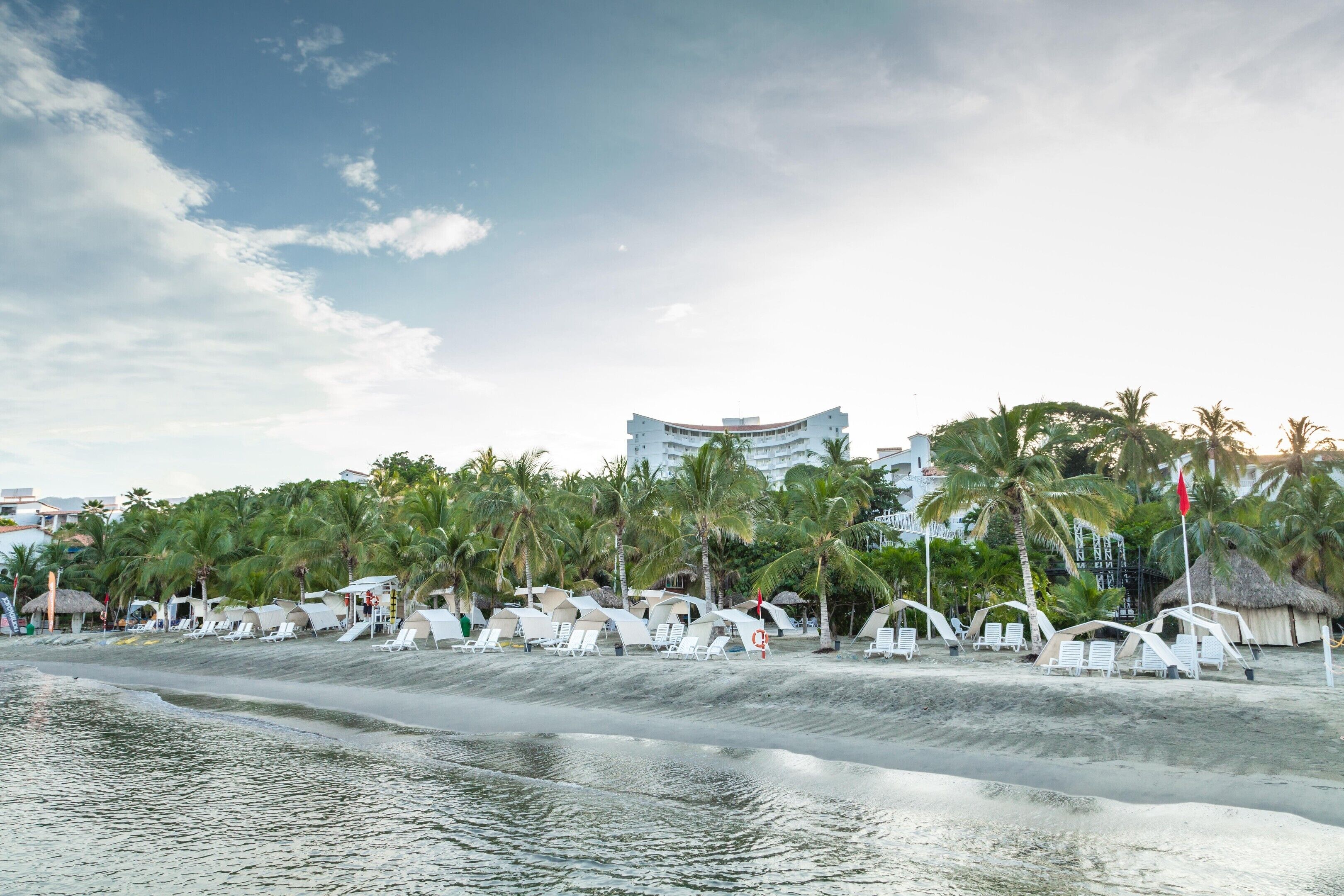 A beach with white tents and lounge chairs, palm trees, and a building in the background.