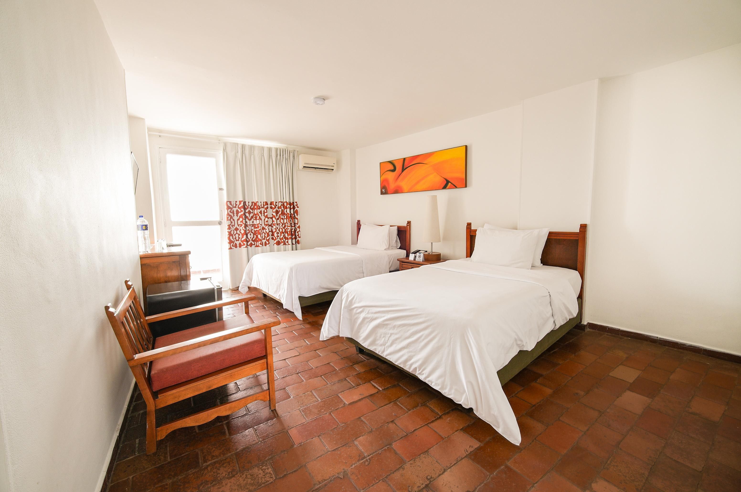 A hotel room with two beds, a desk, a chair, a painting on the wall, and an air conditioning unit.