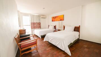 A hotel room with two beds, a desk, a chair, a painting on the wall, and an air conditioning unit.