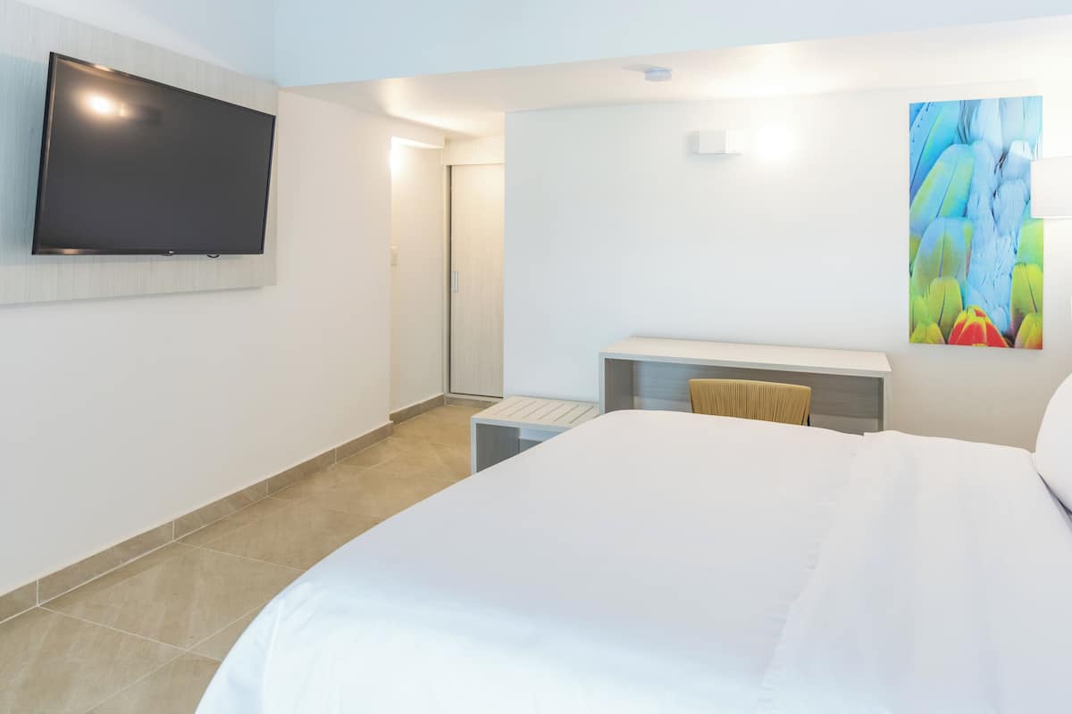 Hotellihuone, jossa on suuri sänky, seinälle asennettu taulutelevisio, pieni työpöytä tuolilla sekä värikäs, abstrakti taulu seinällä.