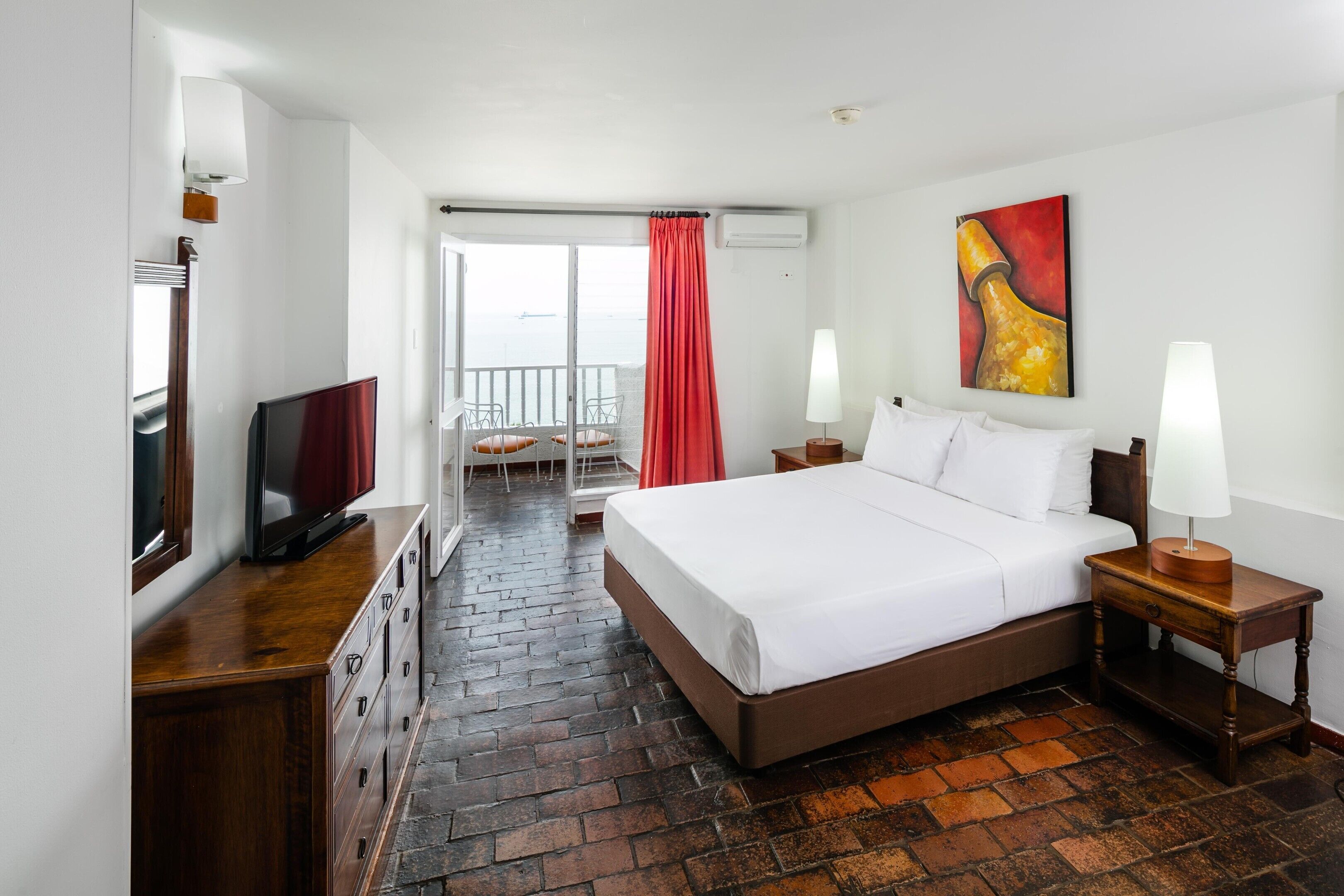 A hotel room with a bed, bedside lamps, a TV, a desk, and a view of the sea through a sliding door.