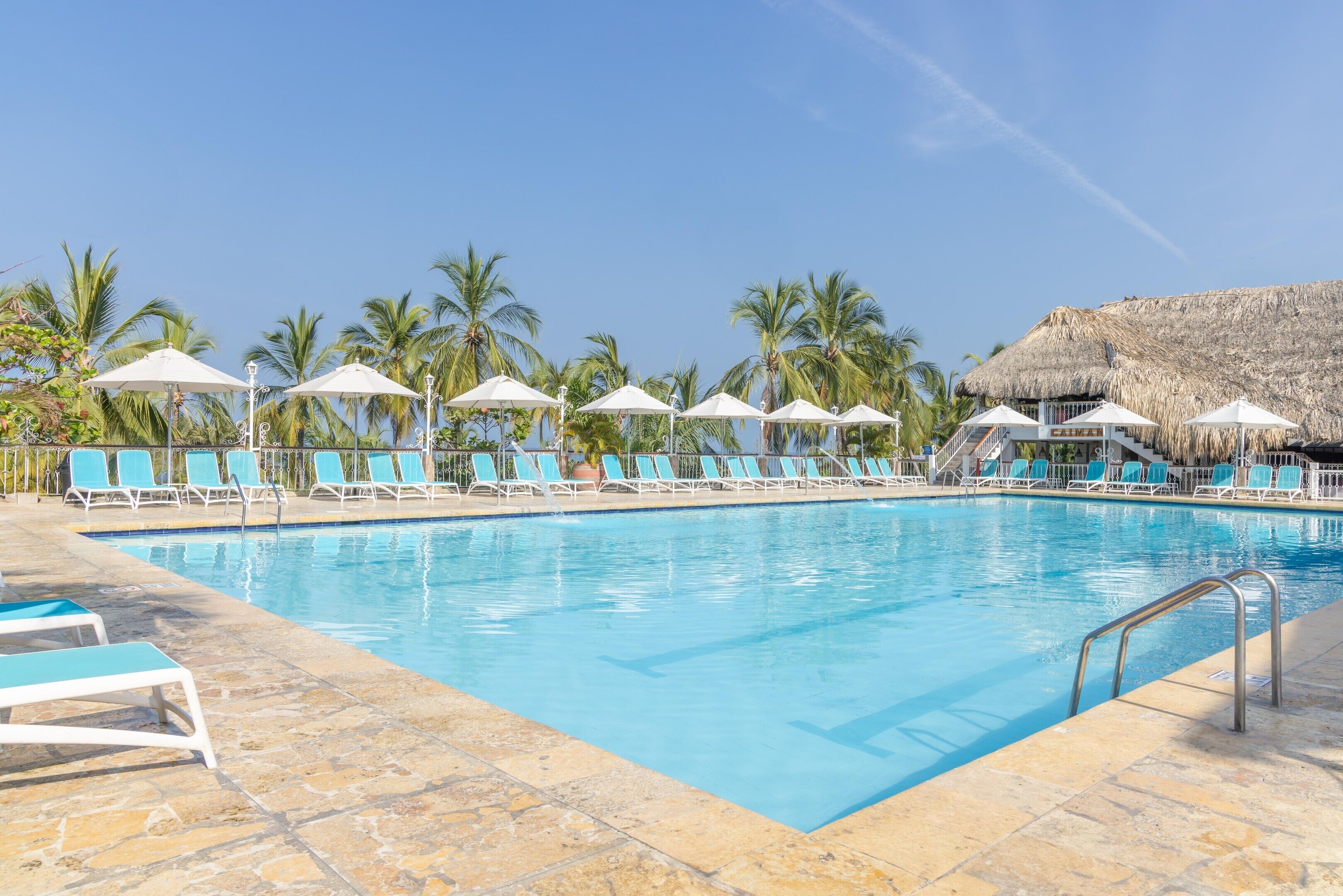 A swimming pool with lounge chairs and umbrellas, surrounded by palm trees and a thatched-roof structure.