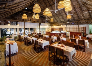 En restaurant med flere borde og stole, et stråtækt tag og hængende fletlamper.