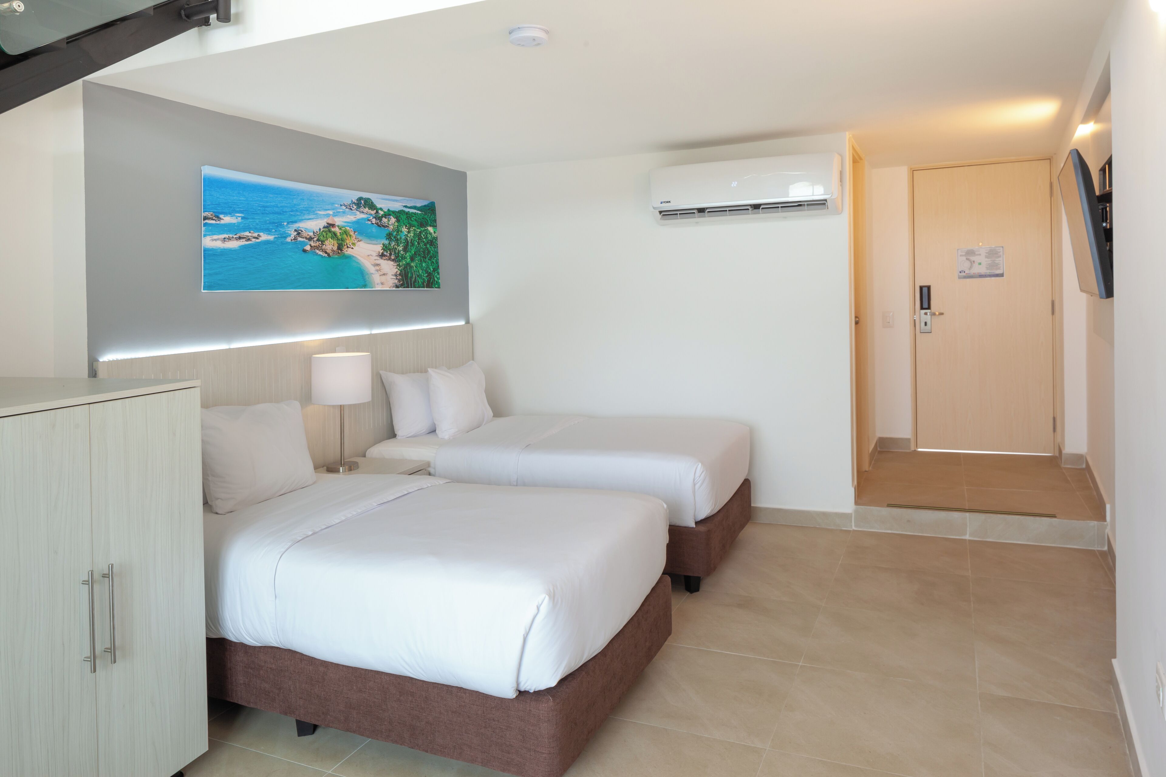 A hotel room with two beds, a desk, a lamp, an air conditioning unit, and a door.