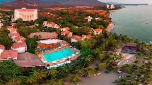 An aerial view of a resort with a swimming pool, beach, and mountain backdrop.
