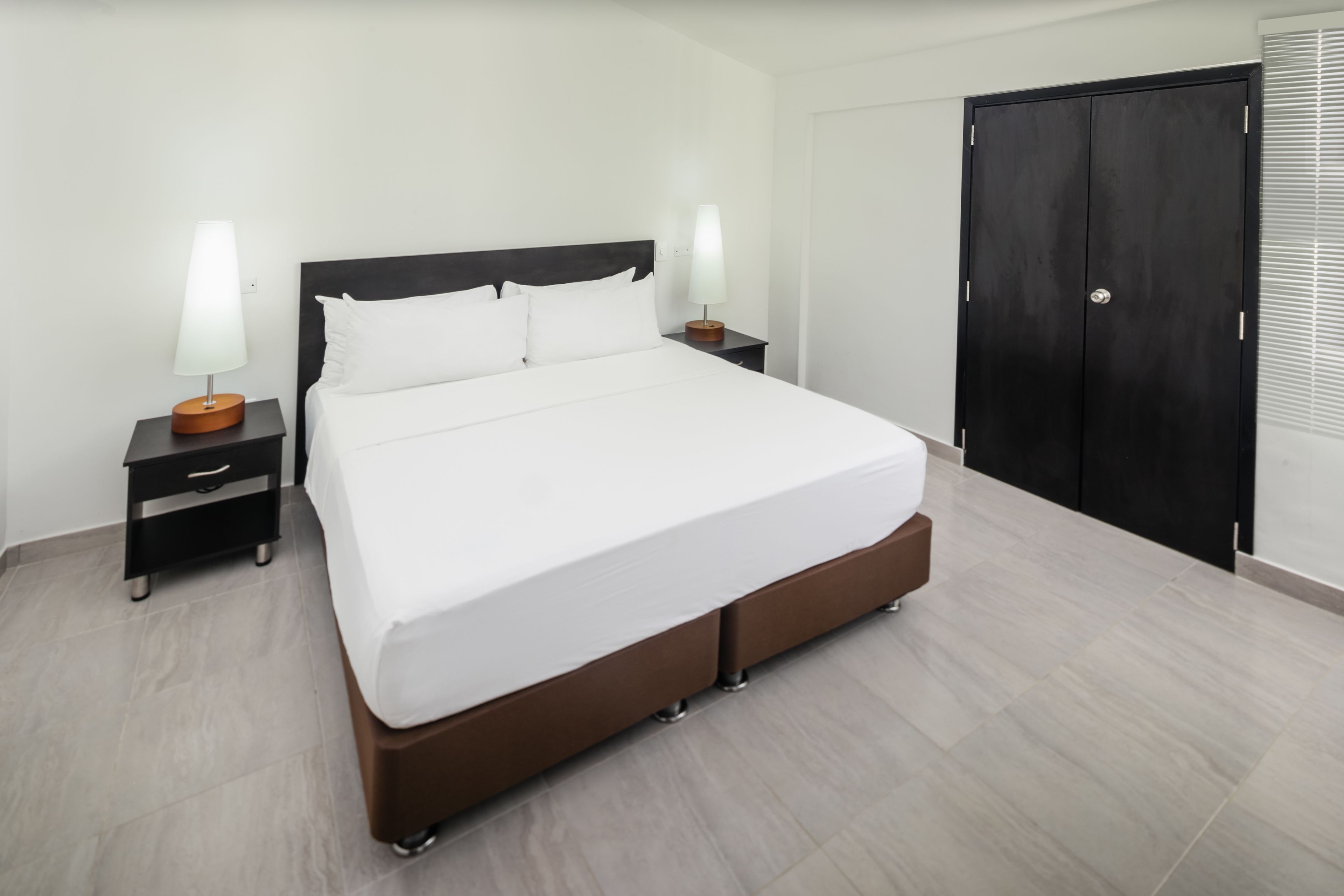 A modern hotel room with a large bed, two bedside tables with lamps, a dark wardrobe, and a tiled floor.