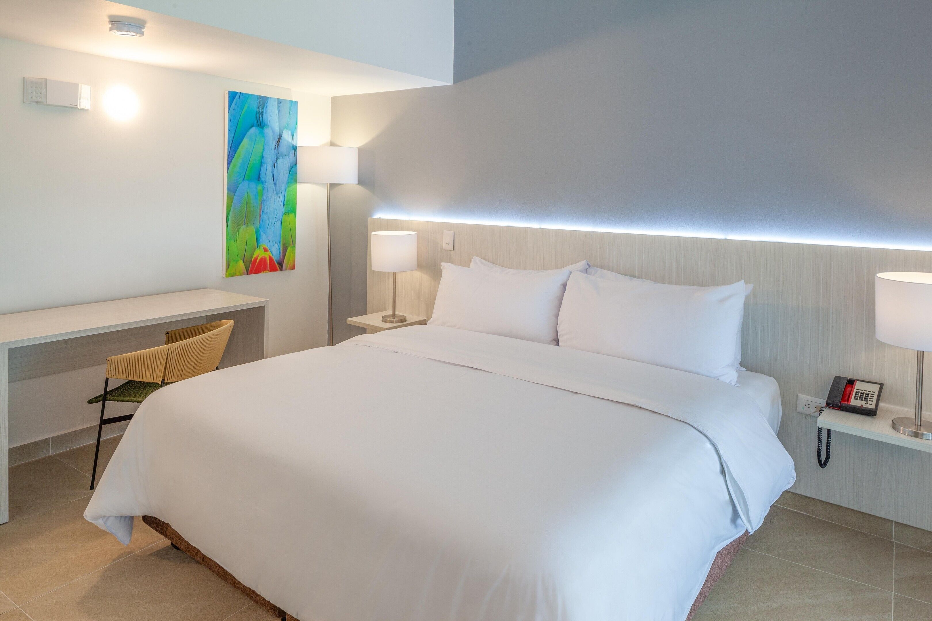 A hotel room with a bed, a desk, a chair, a nightstand with a lamp, and a telephone.