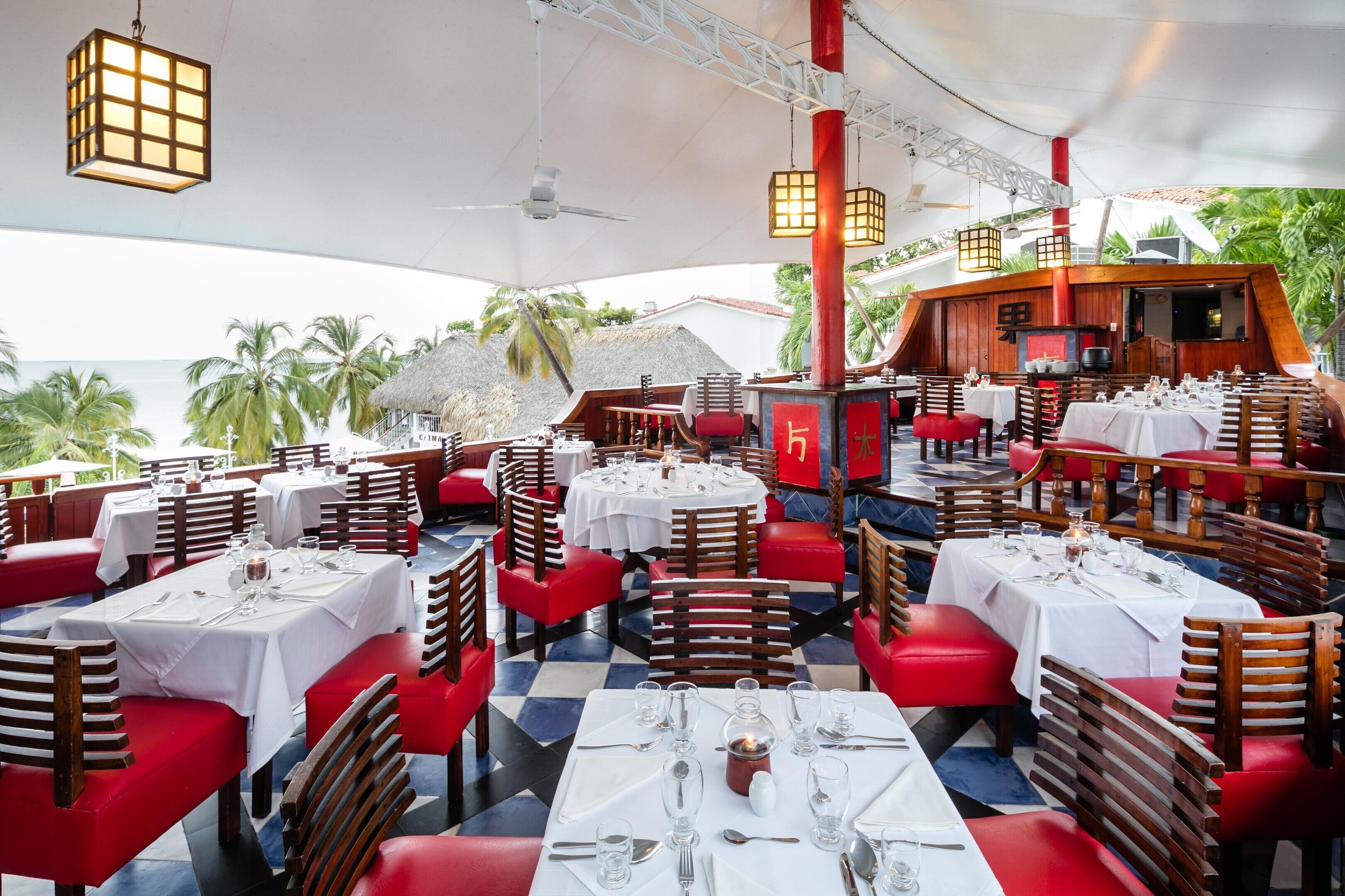 An outdoor restaurant with multiple tables and chairs, a ceiling fan, and a view of the ocean.