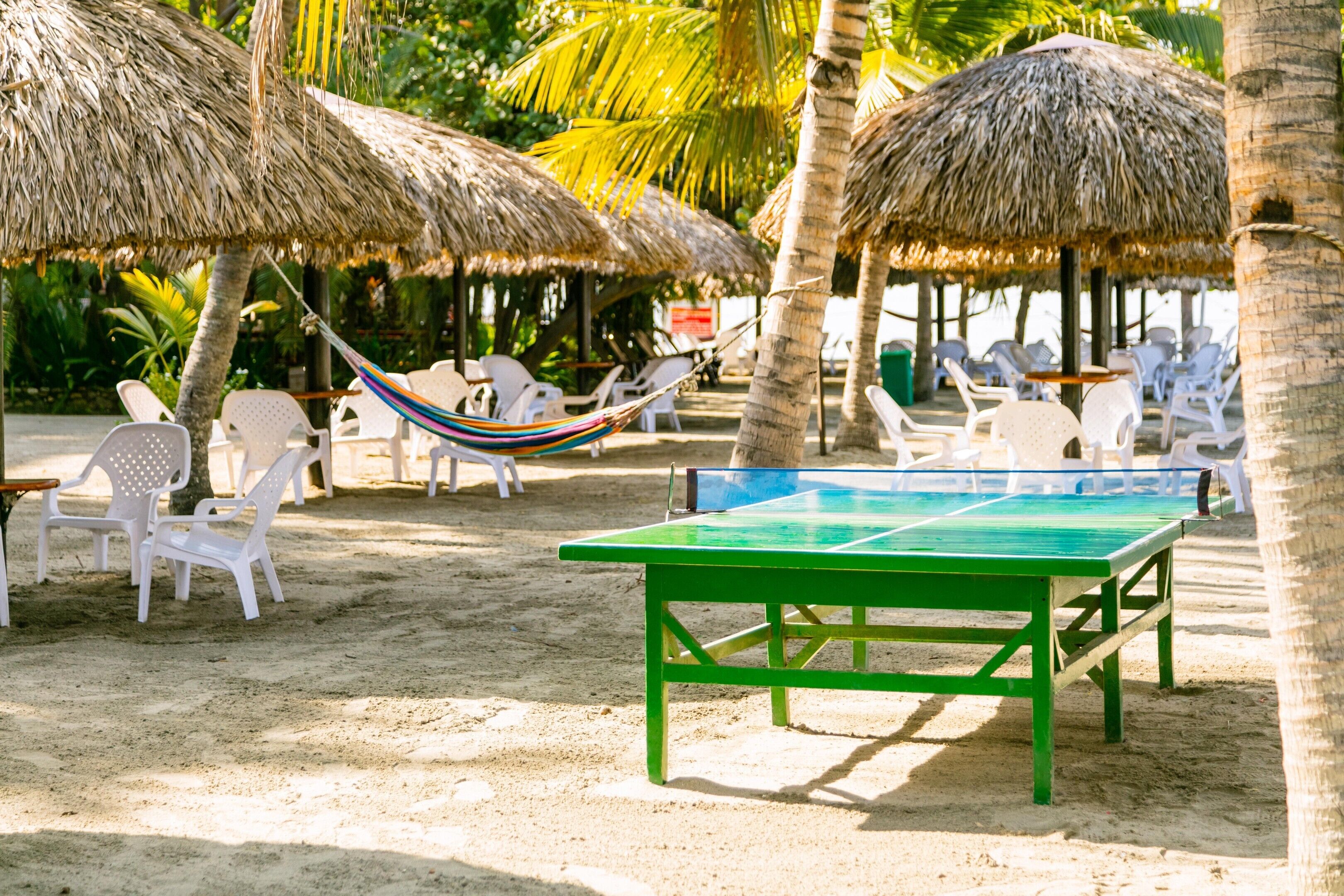 Un paisaje de playa con mesa de ping pong, sillas de plástico blanco y una hamaca.