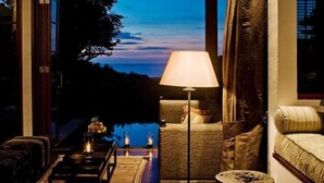 A cozy living room with a sofa, a coffee table, and a lamp. There is a view of the outdoors through a large window. - The Damai (Sukasada)