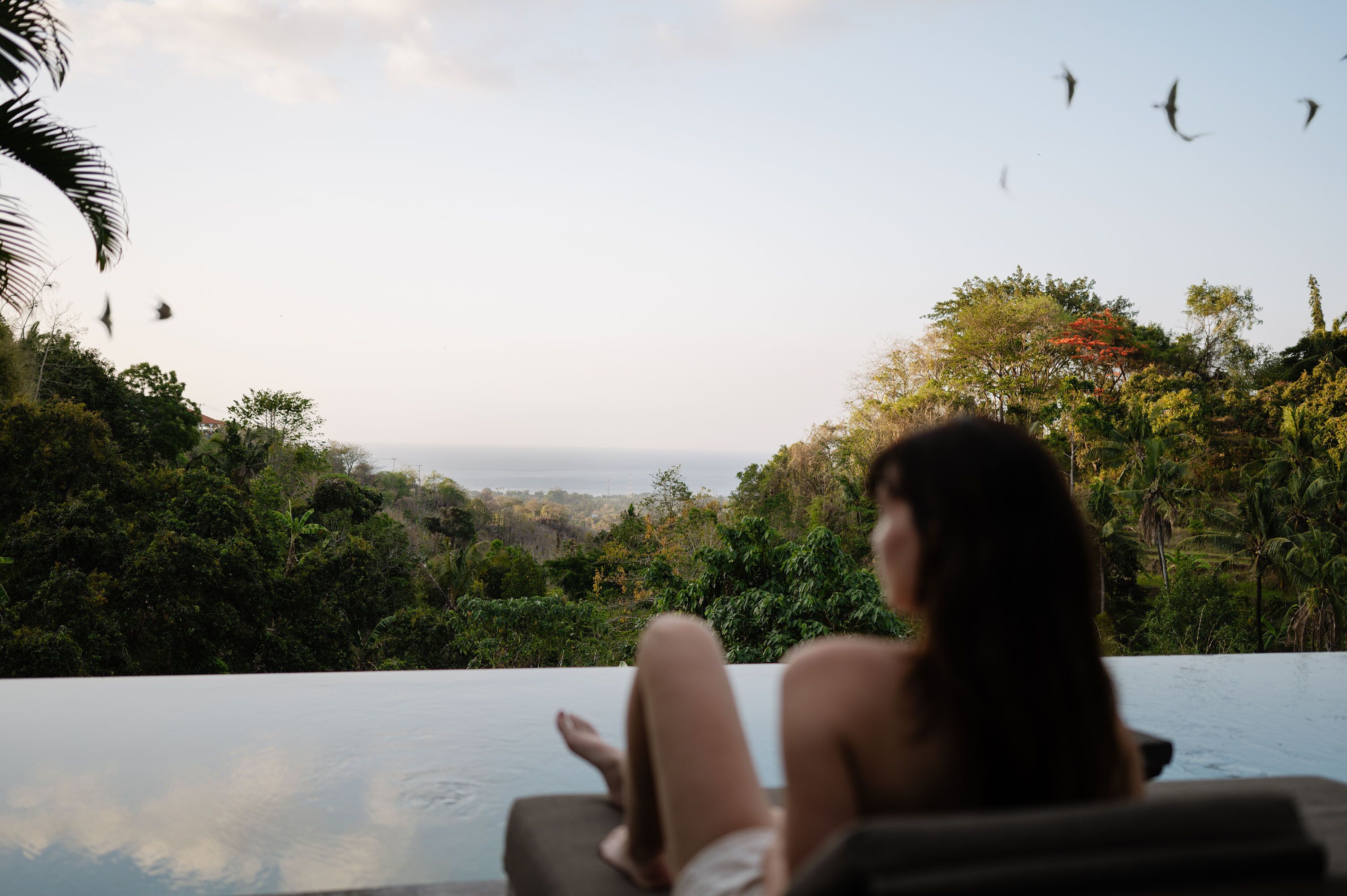 Uma pessoa relaxando junto a uma piscina infinita, com vista para uma paisagem exuberante e o mar.