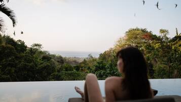 Uma pessoa relaxando junto a uma piscina infinita, com vista para uma paisagem exuberante e o mar.
