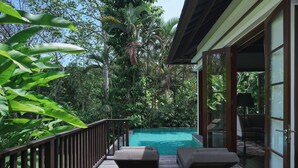 A wooden deck with two lounge chairs overlooking a swimming pool, surrounded by lush tropical vegetation. - The Damai (Sukasada)