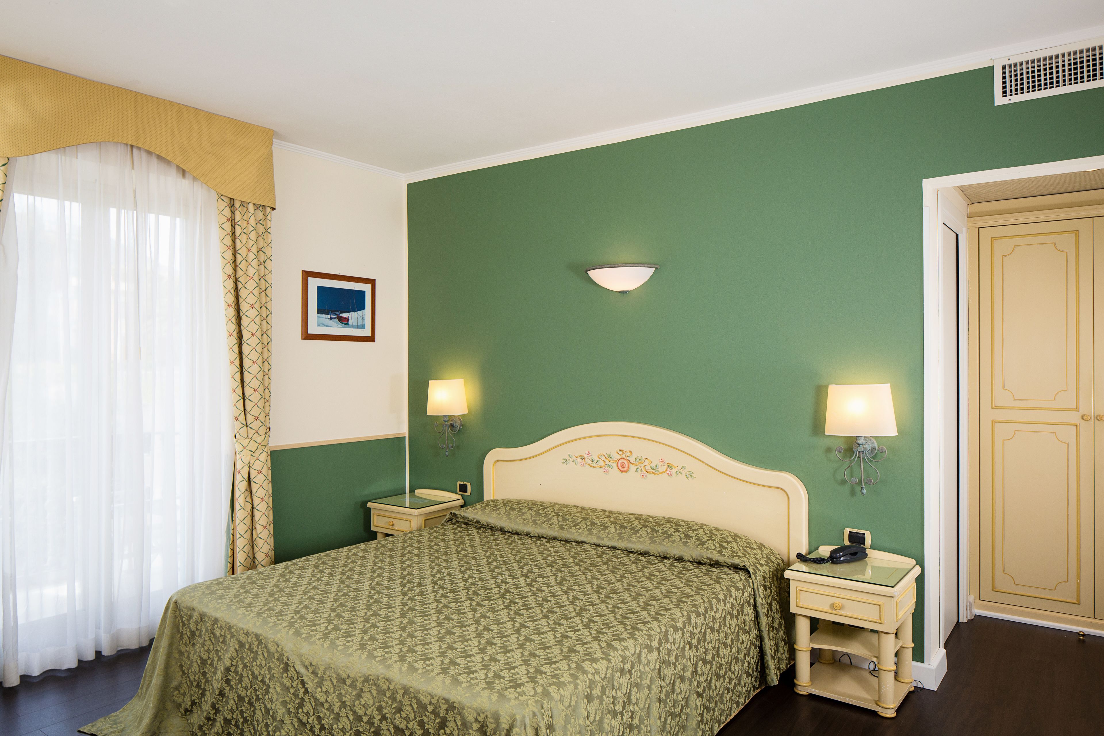 A bedroom with a bed, two bedside tables with lamps, a nightstand, a wall-mounted light fixture, a door, and a window with curtains.