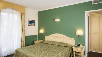 A bedroom with a bed, two bedside tables with lamps, a nightstand, a wall-mounted light fixture, a door, and a window with curtains.