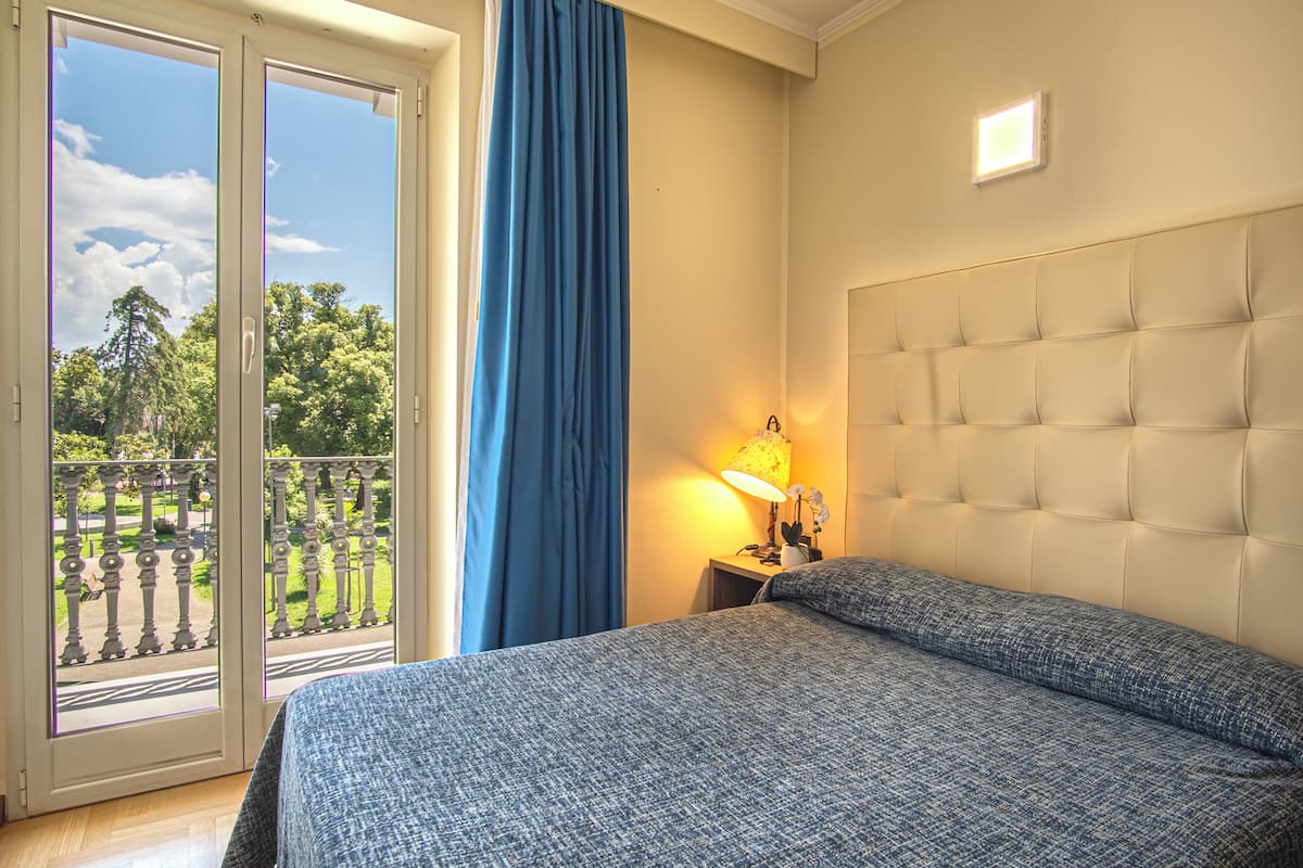A bedroom with a large bed, a nightstand with a lamp, a view of a balcony, and a window with curtains.