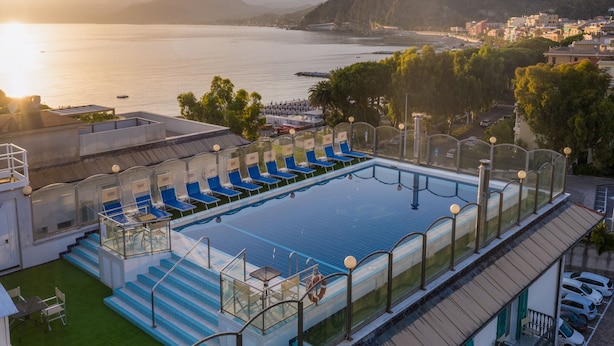 Una piscina en la azotea con sillones azules, barandilla de vidrio y vistas al mar y las colinas.