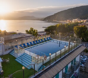 Una piscina en la azotea con sillones azules, barandilla de vidrio y vistas al mar y las colinas.