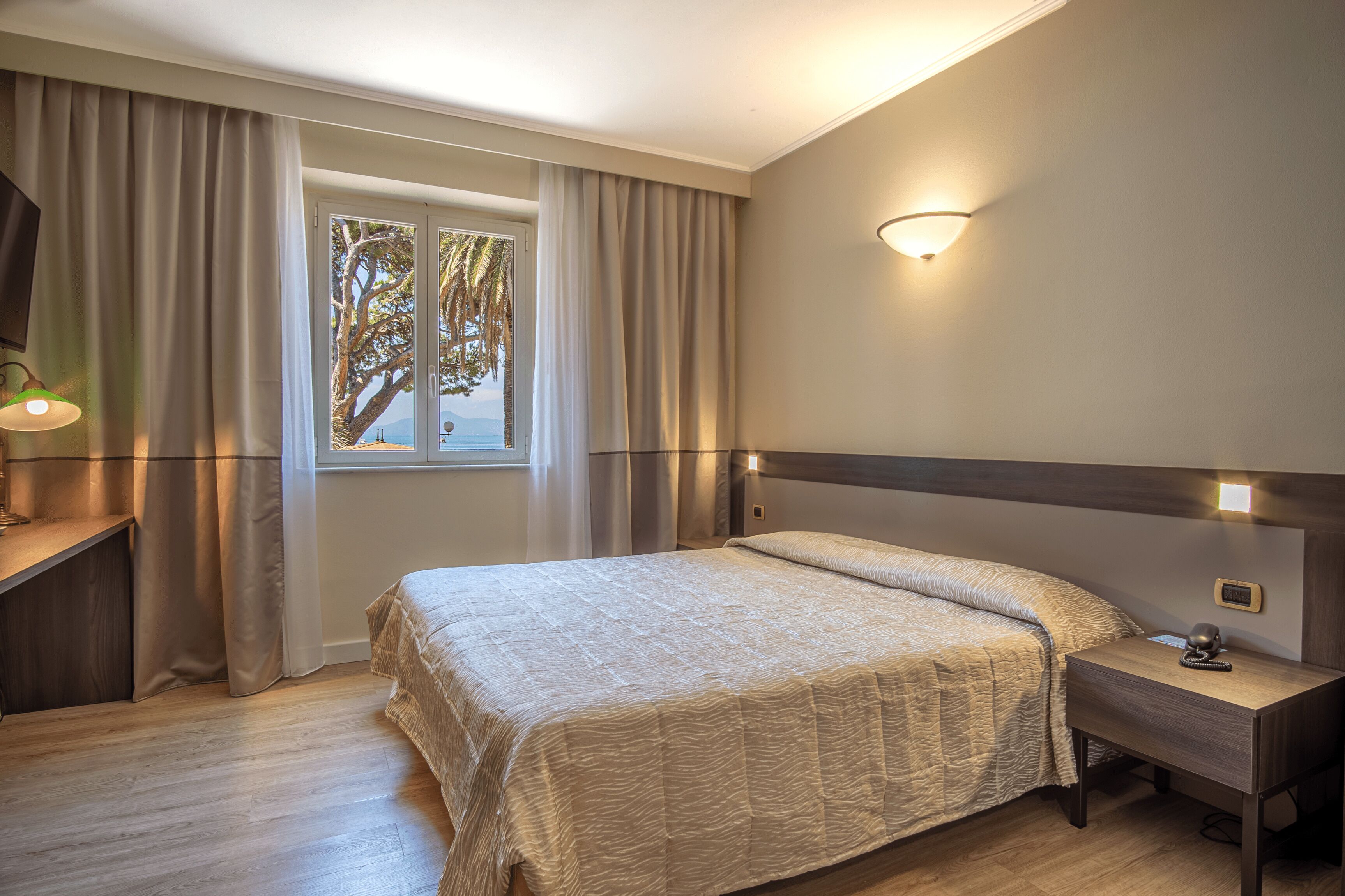 Une chambre à coucher avec un grand lit, une table de chevet, une commode, une fenêtre avec des rideaux et un luminaire fixé au mur.