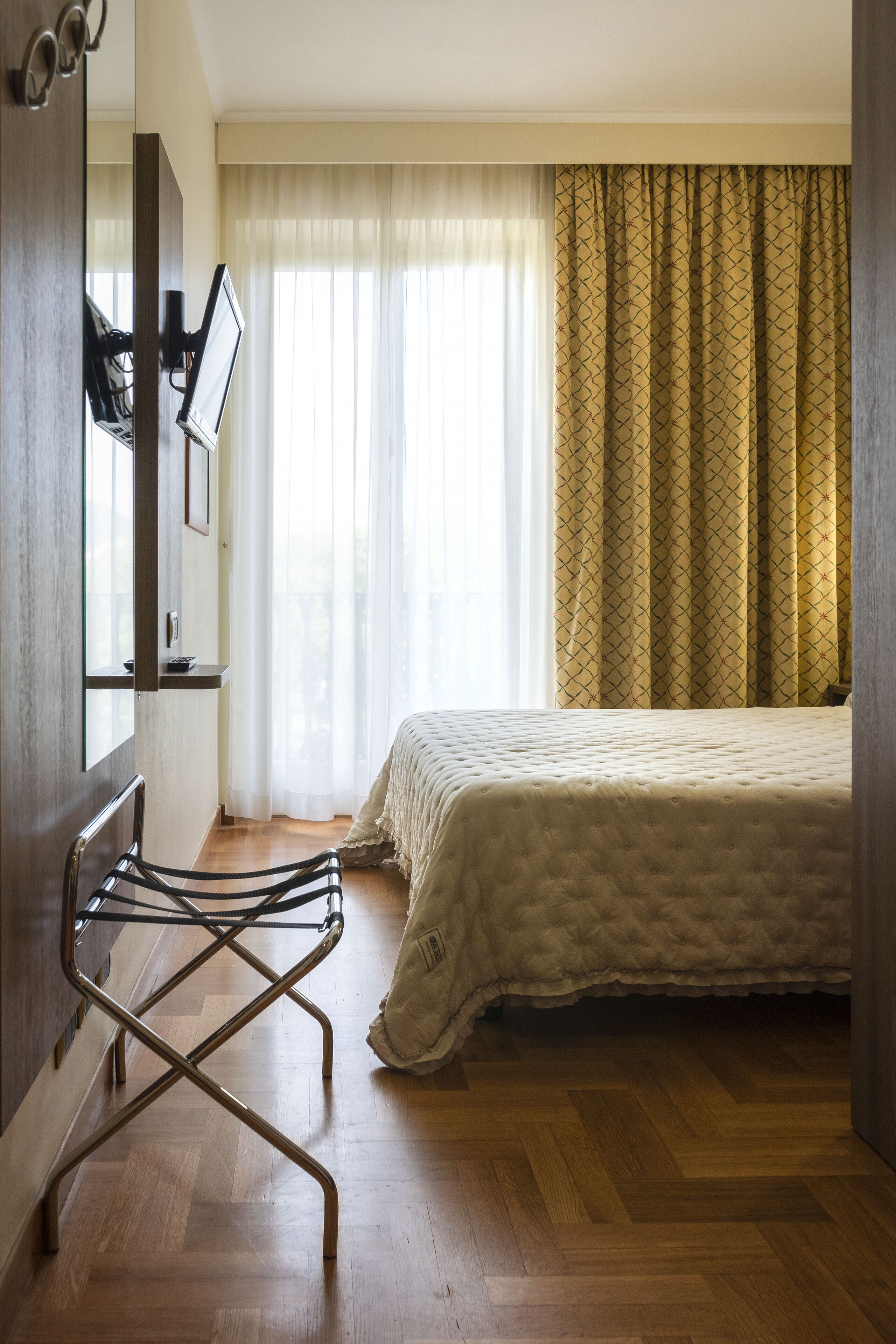 A hotel room with a bed, a chair, a TV mounted on the wall, and a window with curtains.