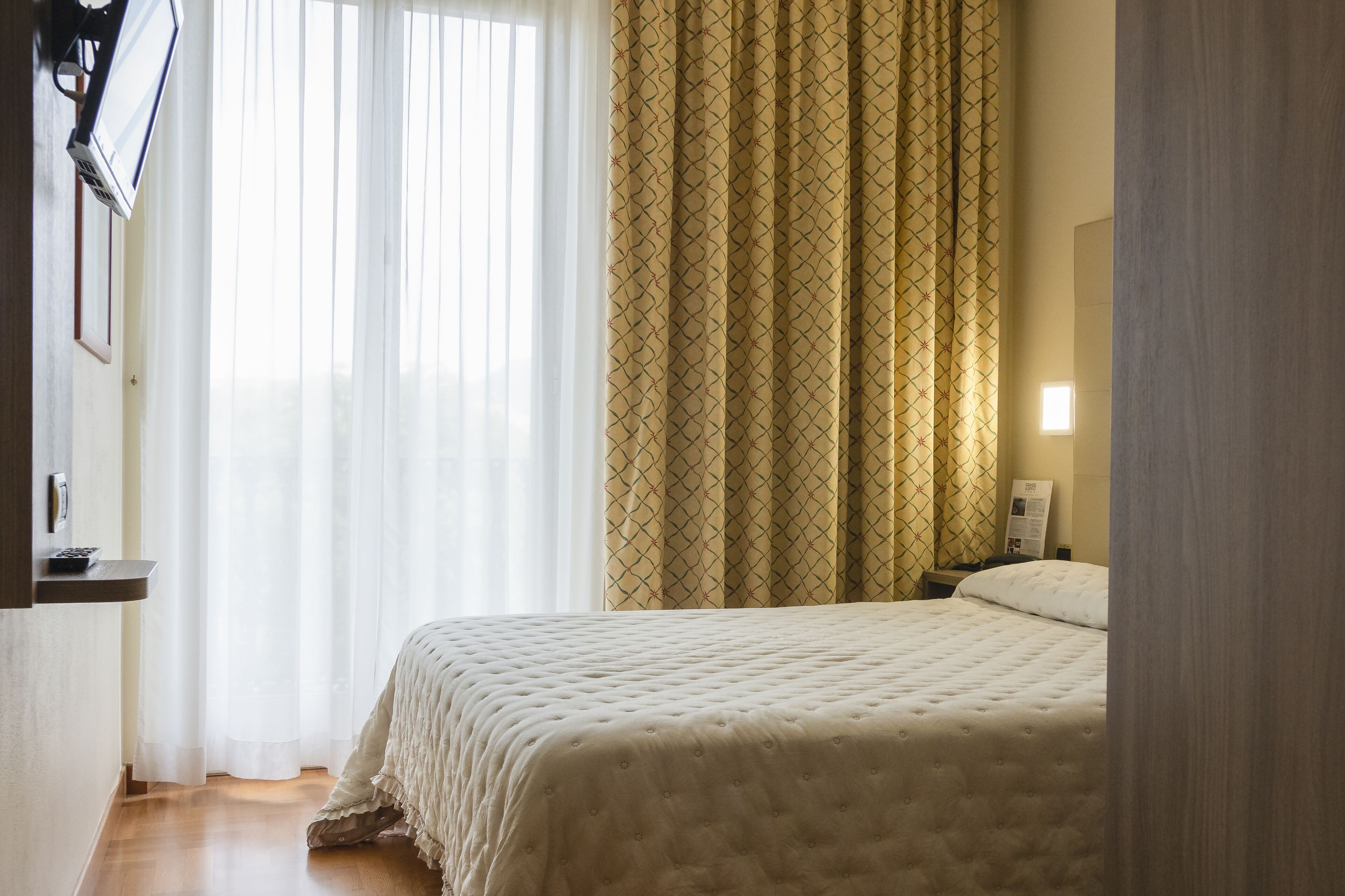 Una camera da letto con un letto, un comodino, una TV a parete e una finestra con tende leggere.