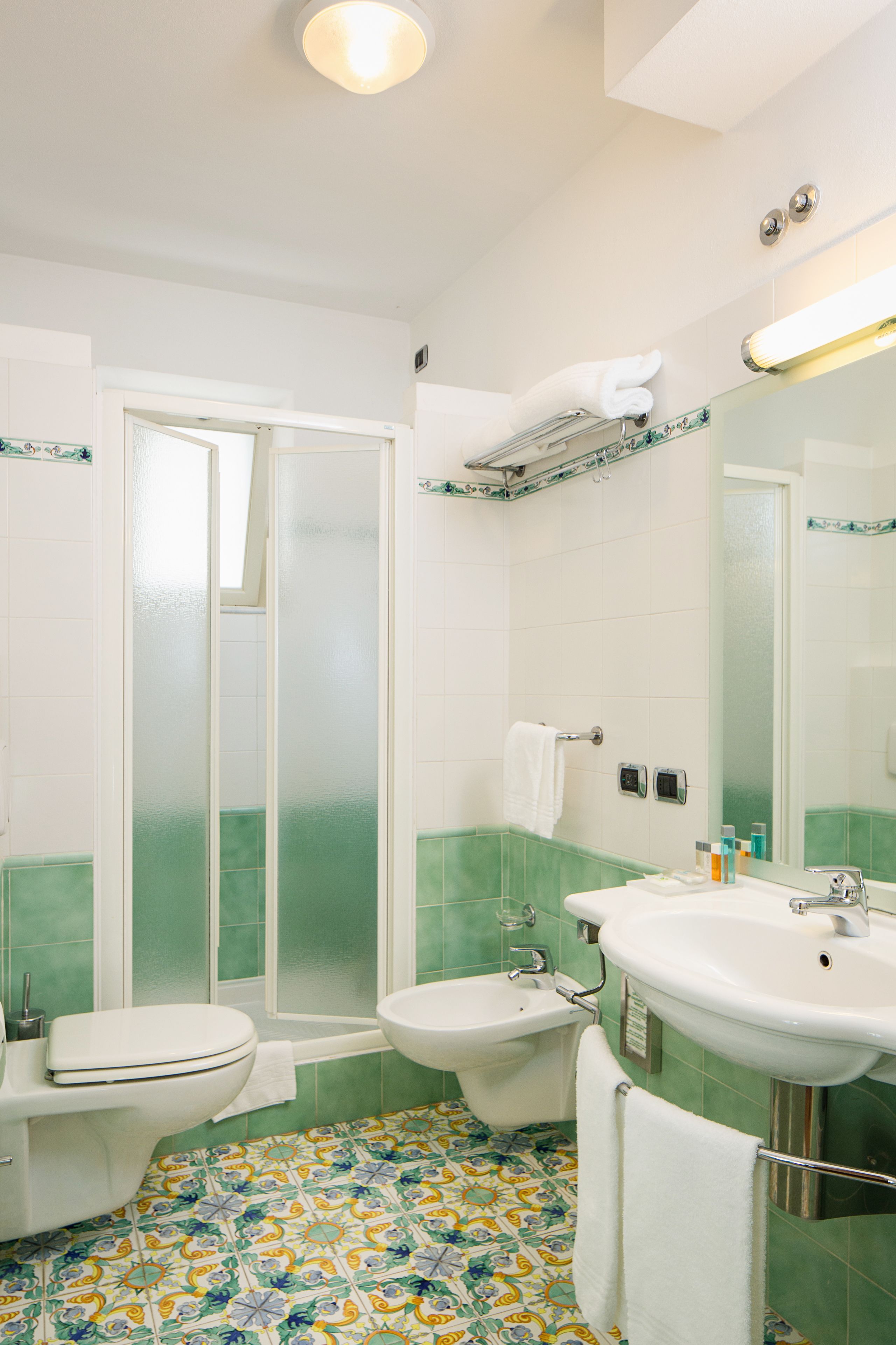 Ein Badezimmer mit Toilette, Bidet und Waschbecken, einer Dusche mit Glastür sowie einem gefliesten Boden mit Blumenmuster.