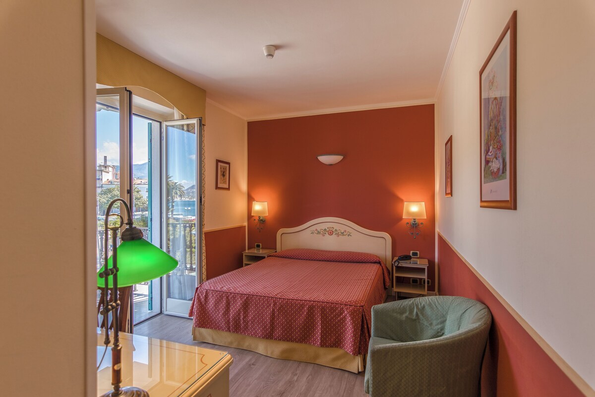 A hotel room with a bed, a chair, a desk, and a balcony with a view.