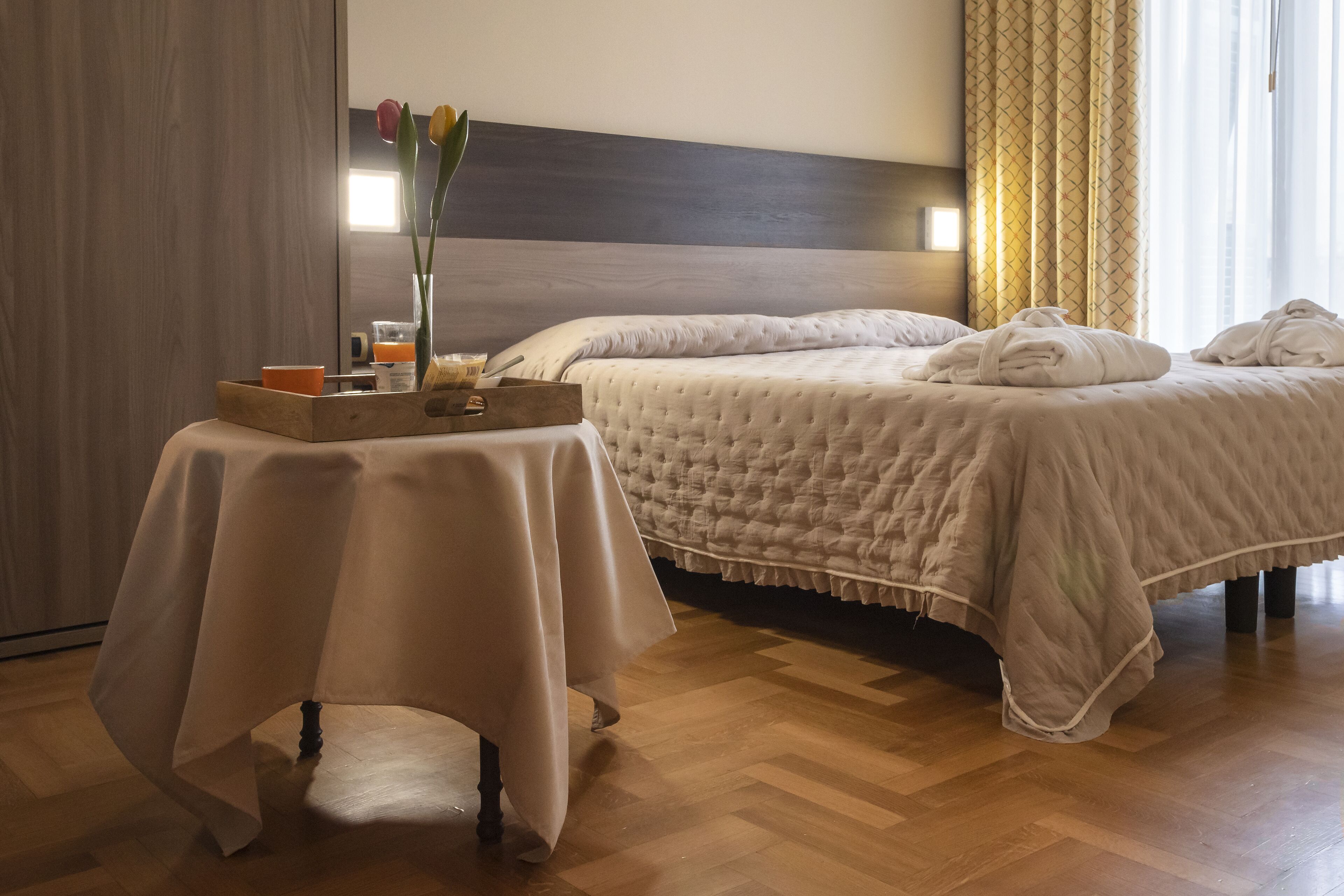 Una habitación de hotel con una cama, una mesita de noche con una bandeja y un florero con flores.