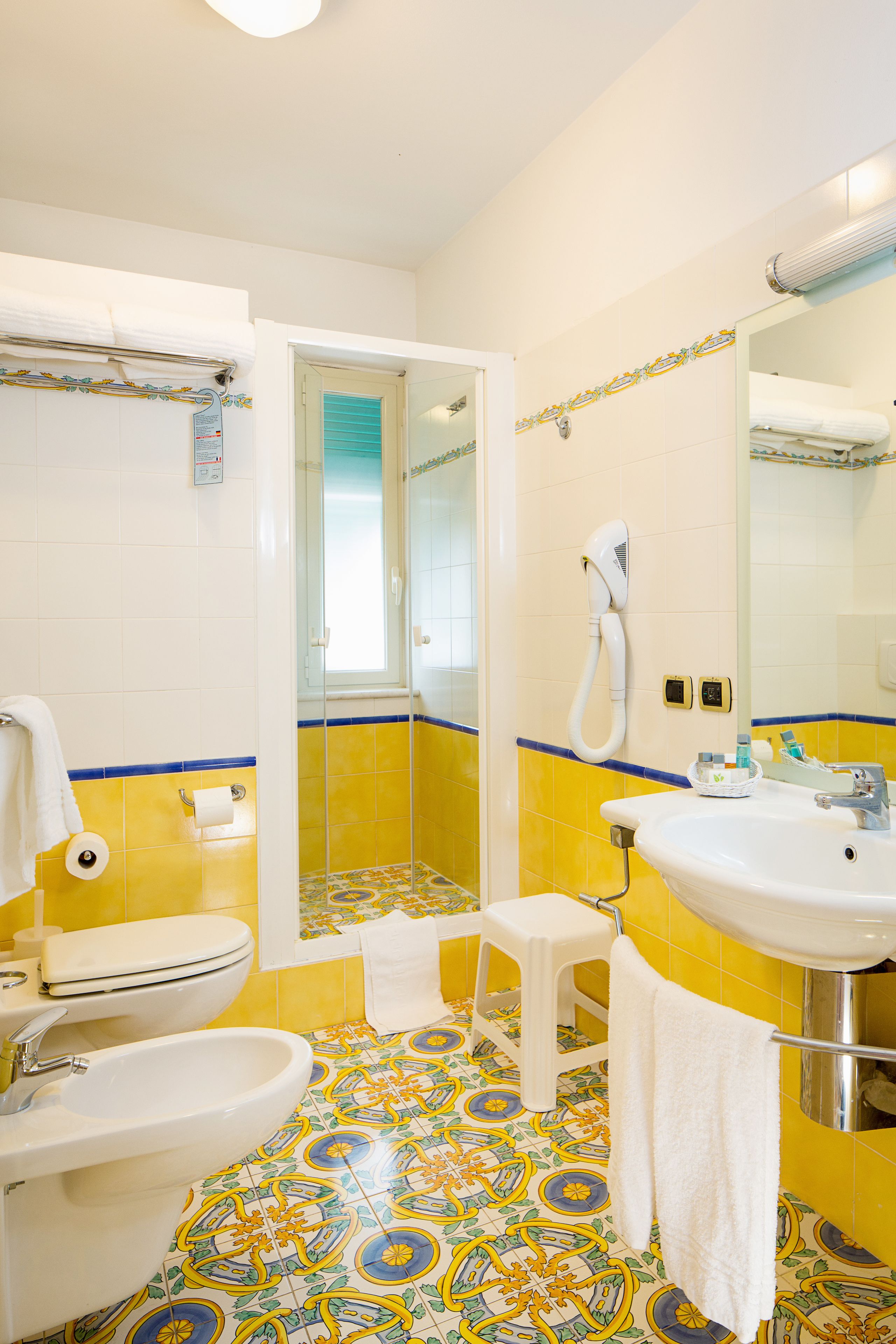 Une salle de bain avec un sol à motifs jaunes et bleus, un lavabo blanc, des toilettes et un espace douche.