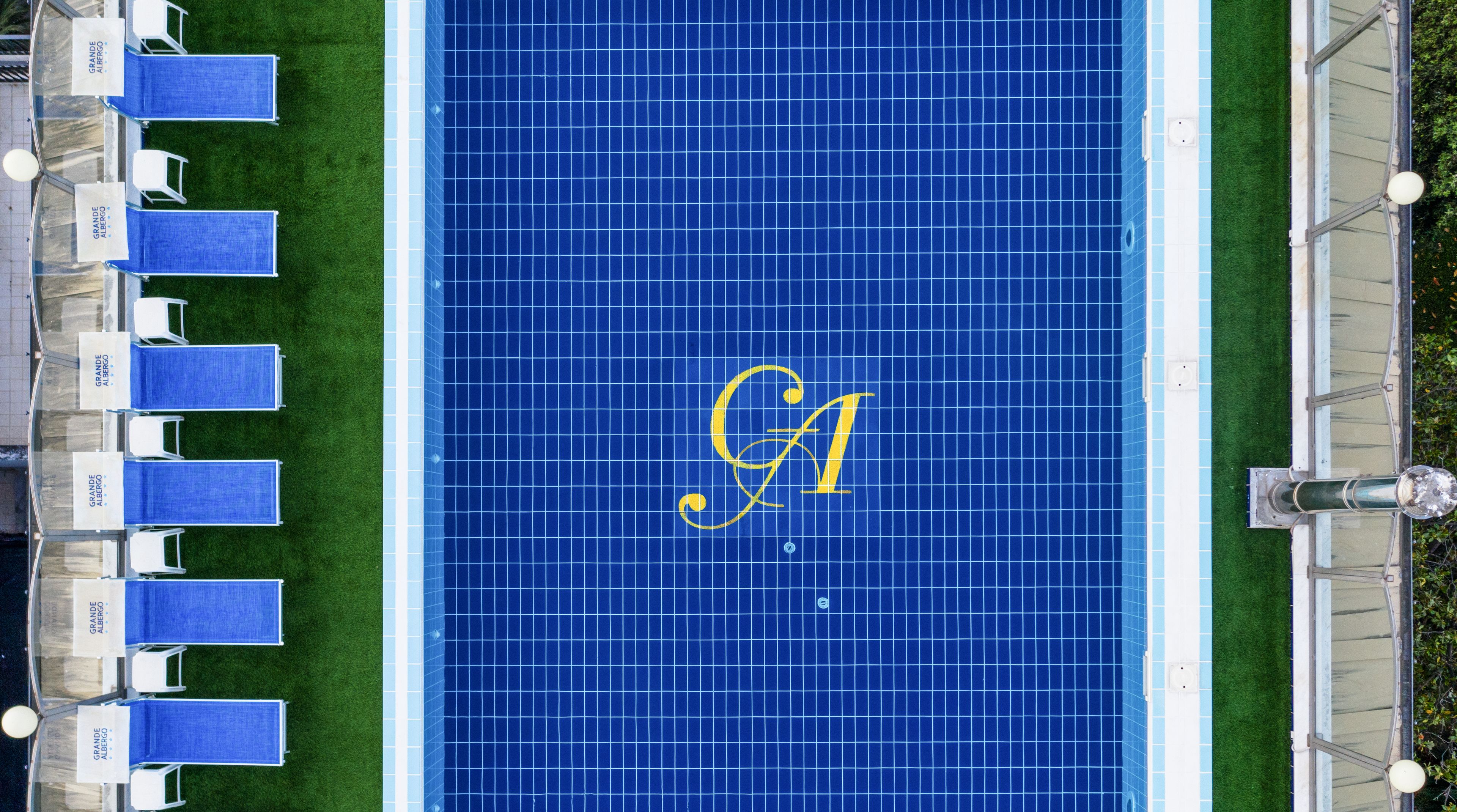 Vue aérienne d'une piscine aux carreaux bleus, avec un grand logo « G » doré au centre.