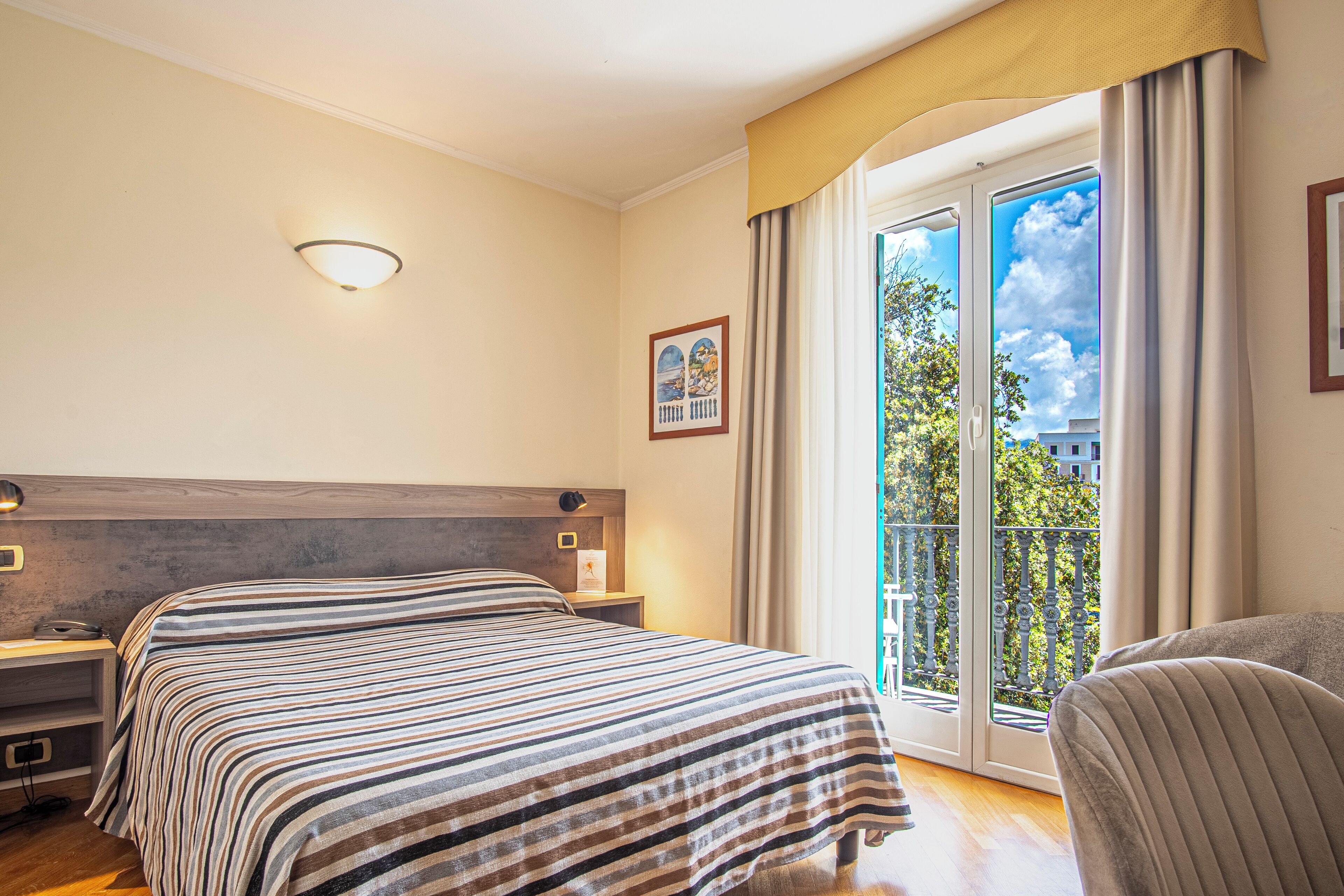 A hotel room with a bed, bedside table, lamp, and a view of a balcony with a railing.