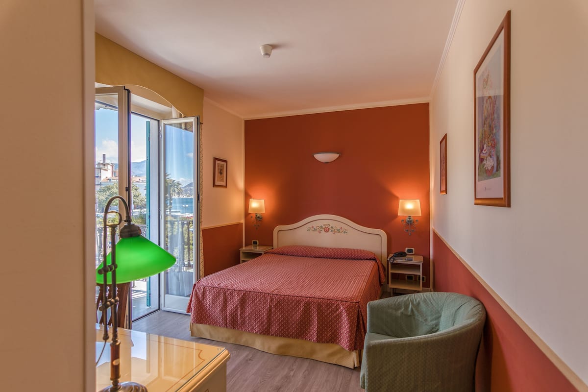 A hotel room with a bed, a chair, a desk, a lamp, and a view of the outside.