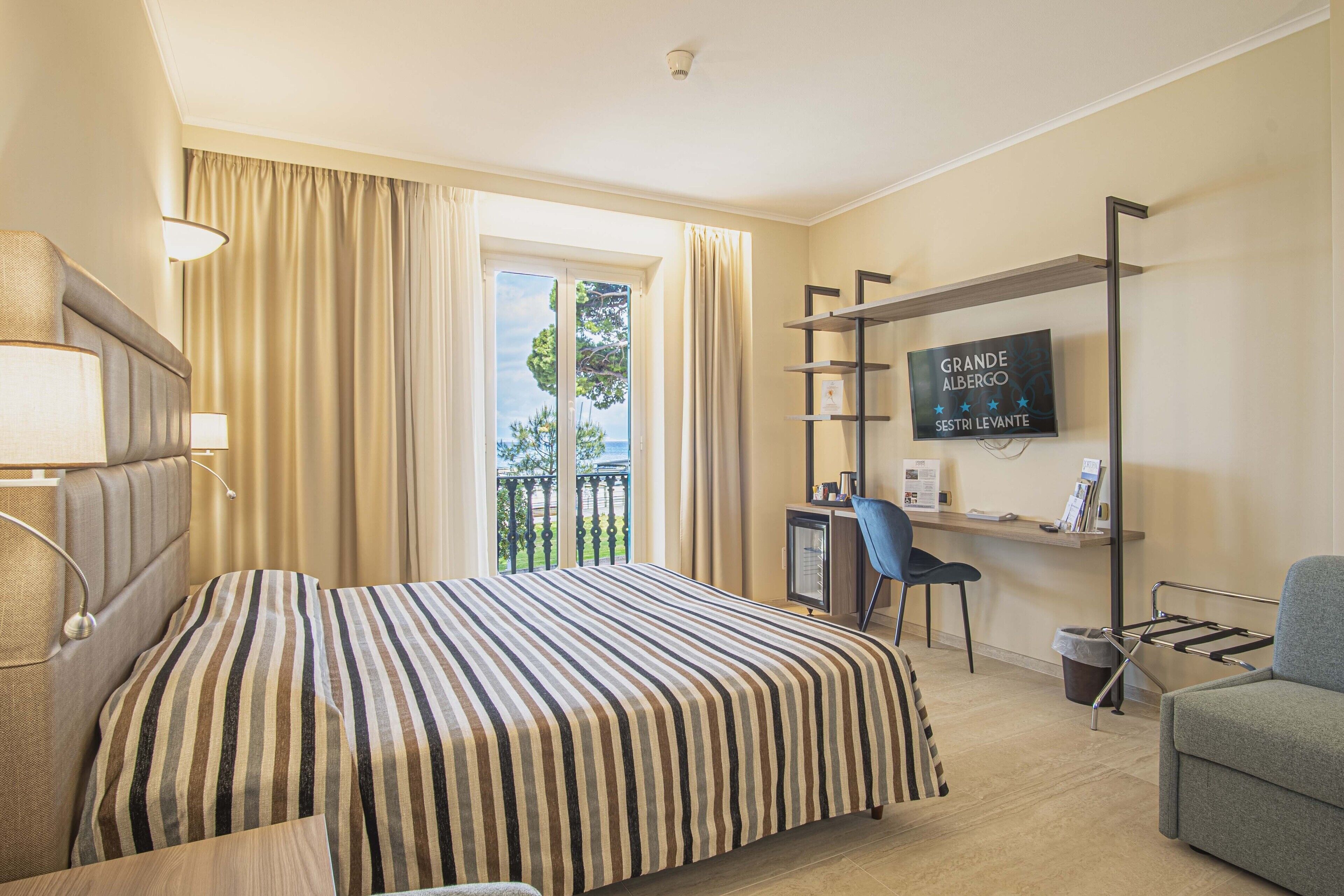 Habitación de hotel con cama, escritorio, silla, televisor y balcón con cortinas.