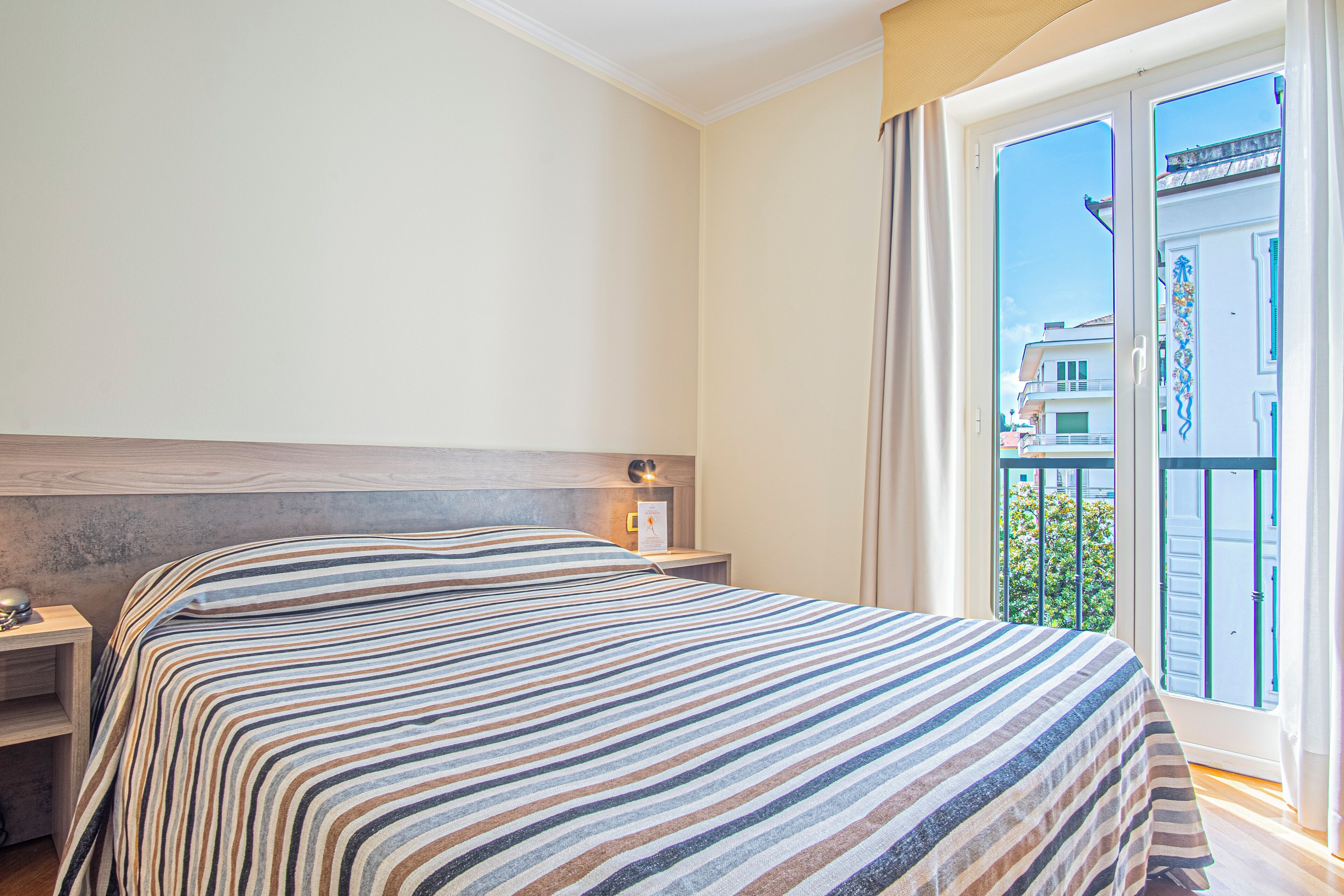 A hotel room with a bed, a nightstand, a window with a view of buildings and greenery, and a balcony.