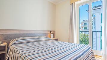 A hotel room with a bed, a nightstand, a window with a view of buildings and greenery, and a balcony.